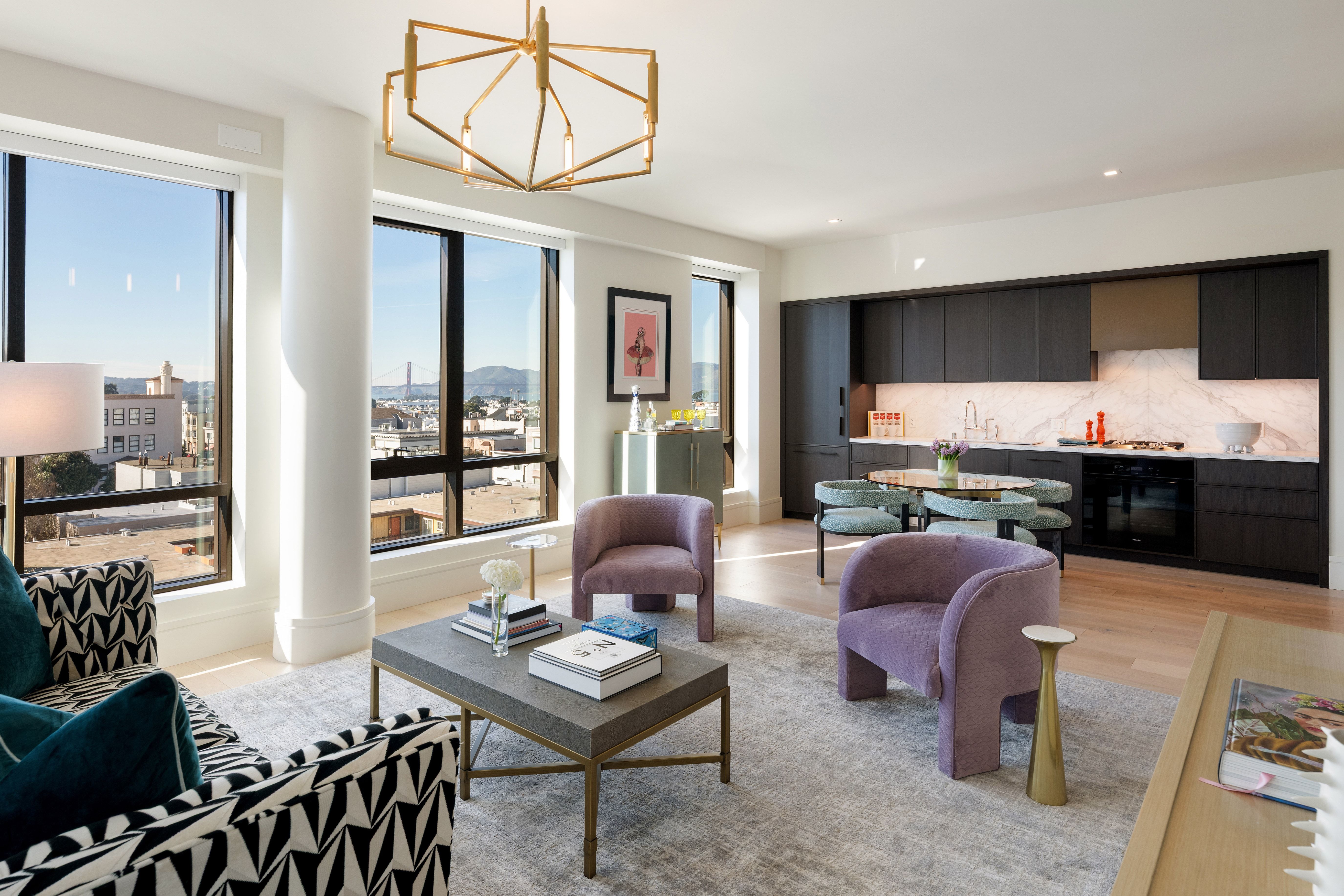 This sophisticated, open-plan living space features a modern aesthetic with a chic lounge area and an integrated dark-toned kitchen. The room is anchored by vibrant purple accent chairs and a stylish geometric patterned sofa, all set against a neutral, light-filled backdrop that frames impressive city views including the Golden Gate Bridge. High-end finishes, such as the sculptural gold chandelier and minimalist marble backsplash, create an elegant and cohesive atmosphere perfect for contemporary urban living.