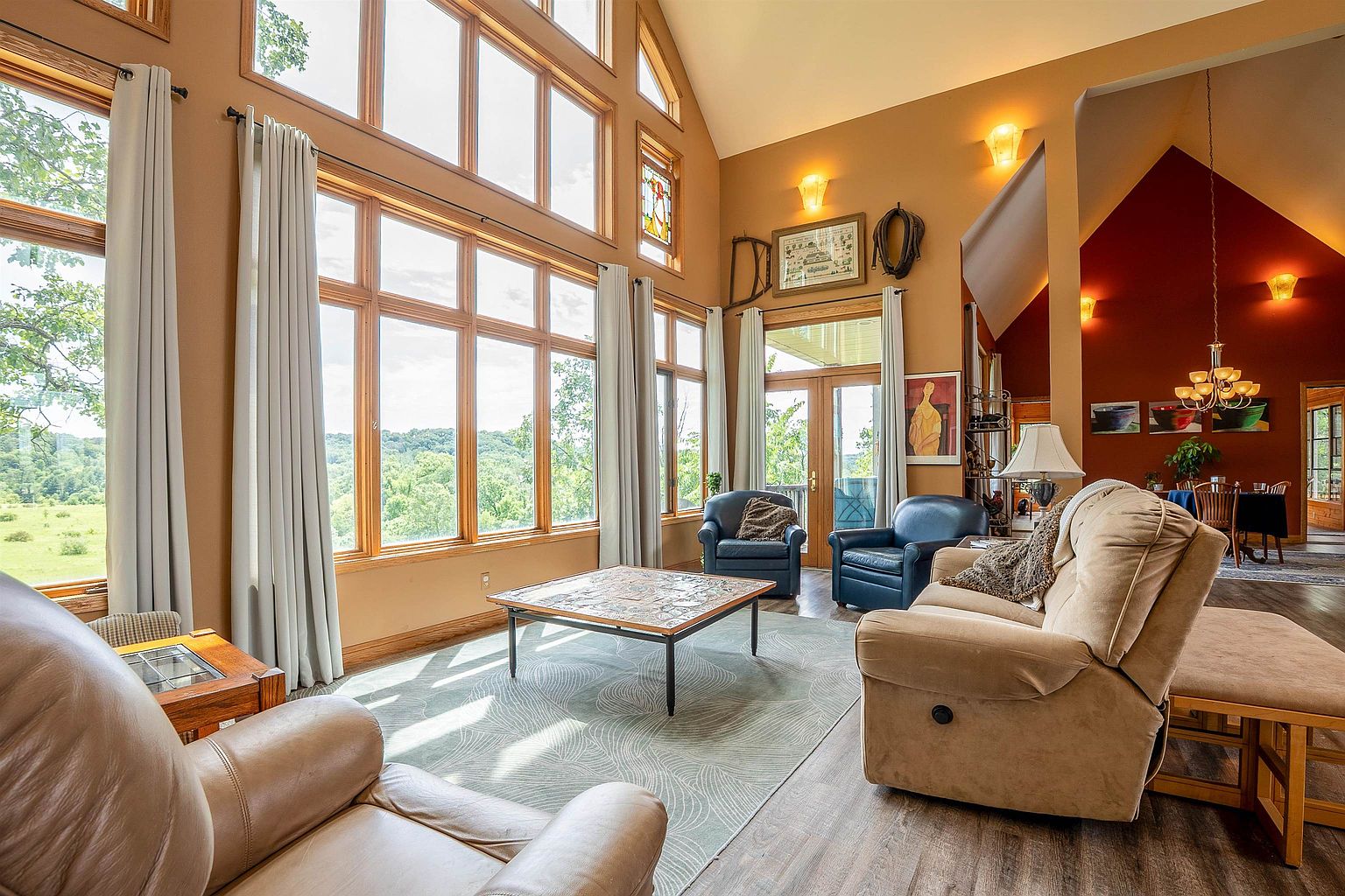 This bright and spacious living room features soaring vaulted ceilings and an expansive wall of windows that provide natural light and scenic views of the surrounding landscape. The space is anchored by a comfortable furniture arrangement including a plush sofa and leather armchairs on a patterned area rug, creating a warm and inviting atmosphere. A combination of earth-toned wall colors and rustic decorative elements gives the room a cozy, lodge-like aesthetic.