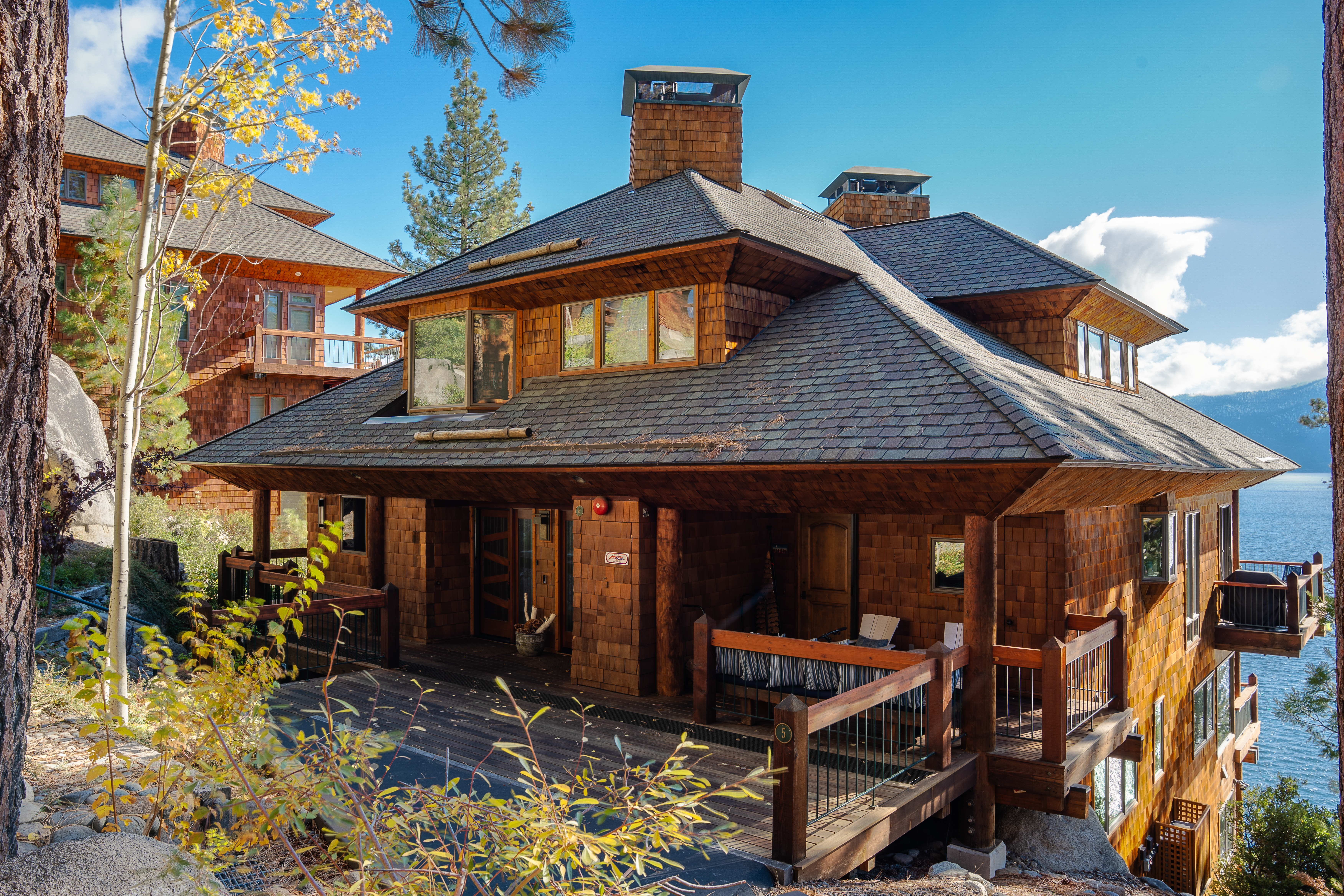 This is a front exterior view of a multi-story, wood-shingled house with a unique architectural design. The house features a complex roofline with multiple gables and dormers, a covered porch with wooden railings, and a partial view of a lake in the background. The surrounding landscape includes trees and foliage, adding to the property's natural appeal.