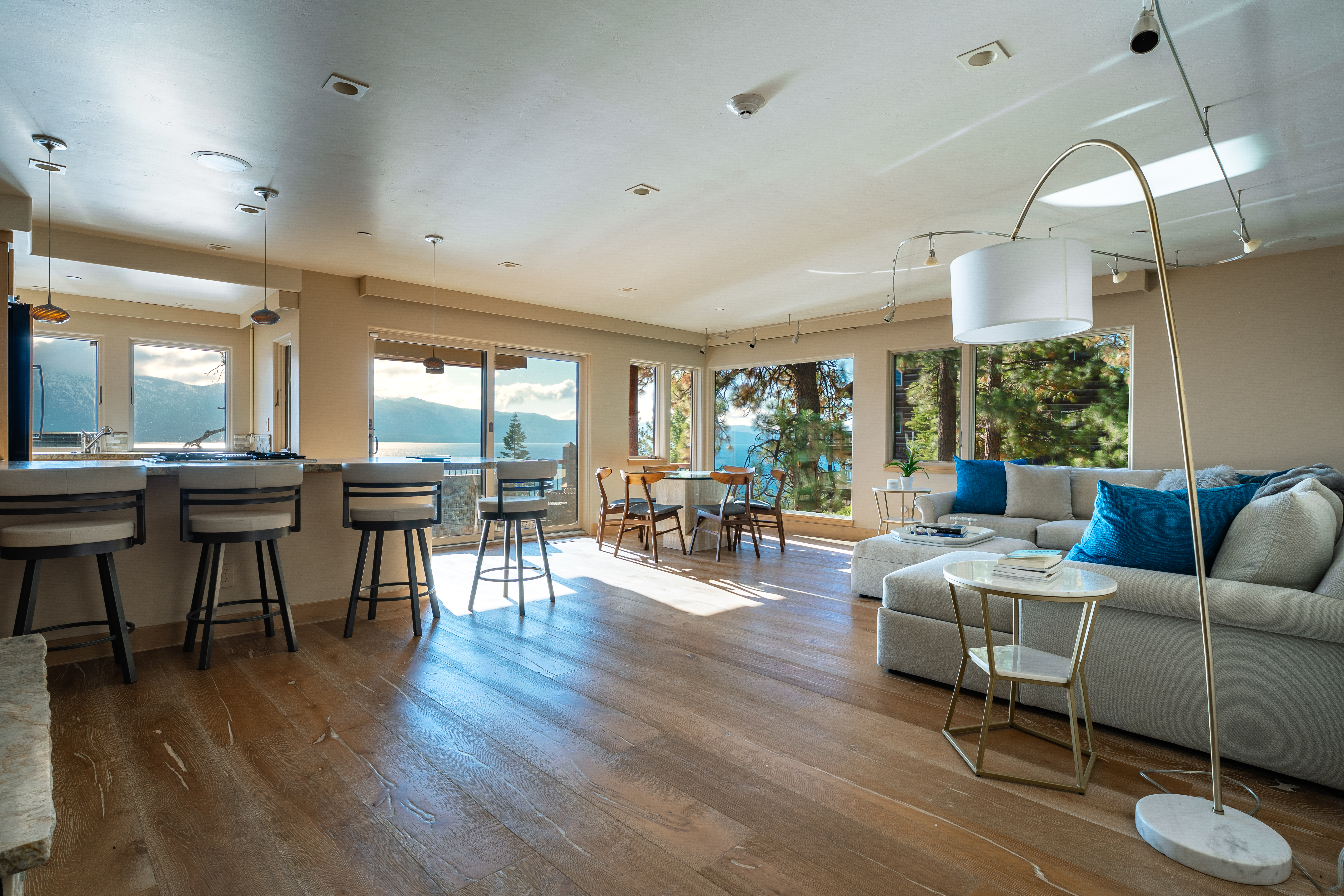 This is a bright and airy living room featuring hardwood floors, a large sectional sofa with blue accent pillows, and floor-to-ceiling windows offering stunning lake views. The open-concept design seamlessly connects the living area to the kitchen and dining space, creating a spacious and inviting atmosphere. The room is well-lit with a combination of natural light and modern lighting fixtures.