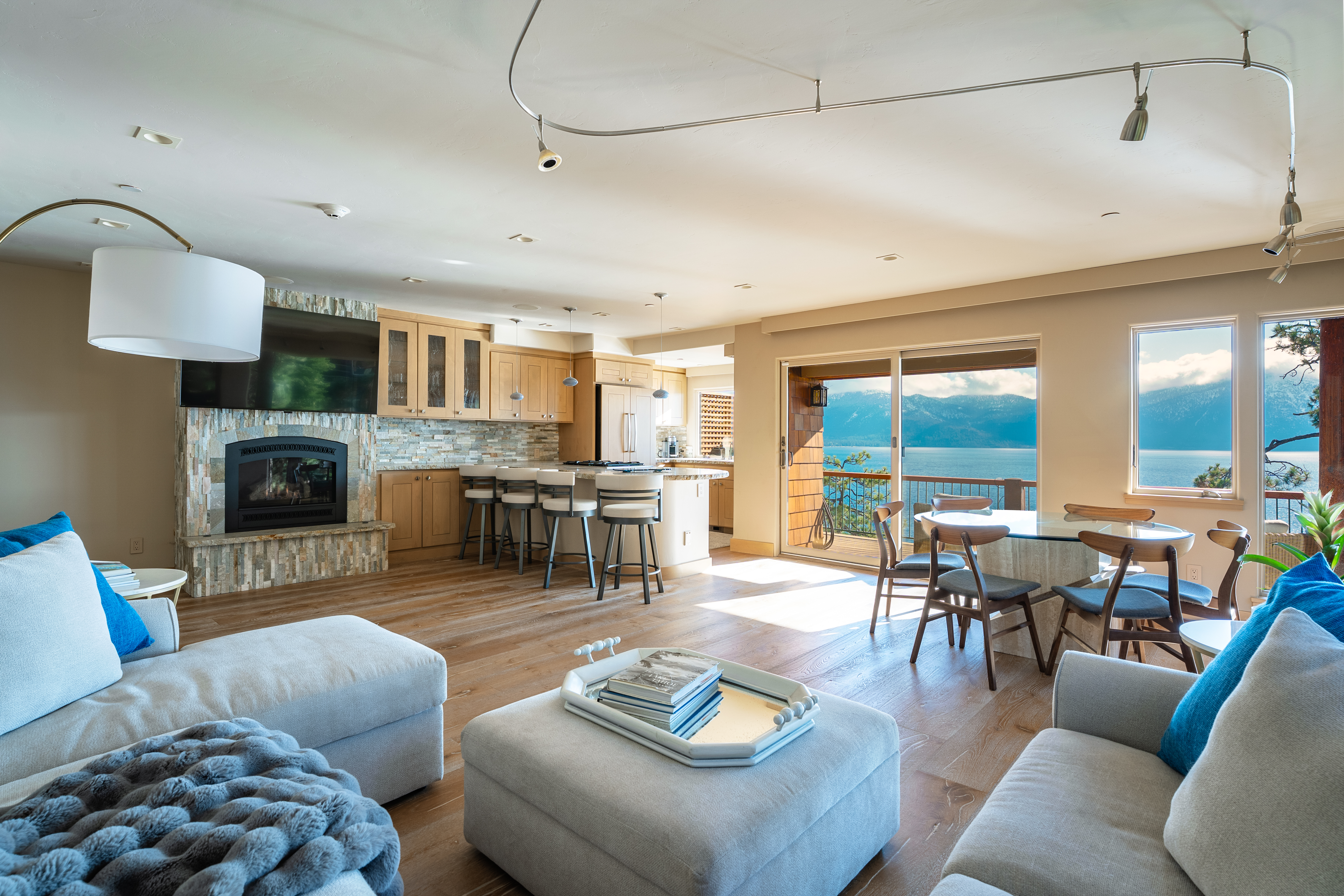 This is an interior shot of a combined living room, dining area, and kitchen, showcasing an open-concept design. The living room features comfortable seating and a fireplace, while the dining area offers a view of the water. The kitchen is equipped with modern appliances and a breakfast bar, creating a seamless flow between the spaces.
