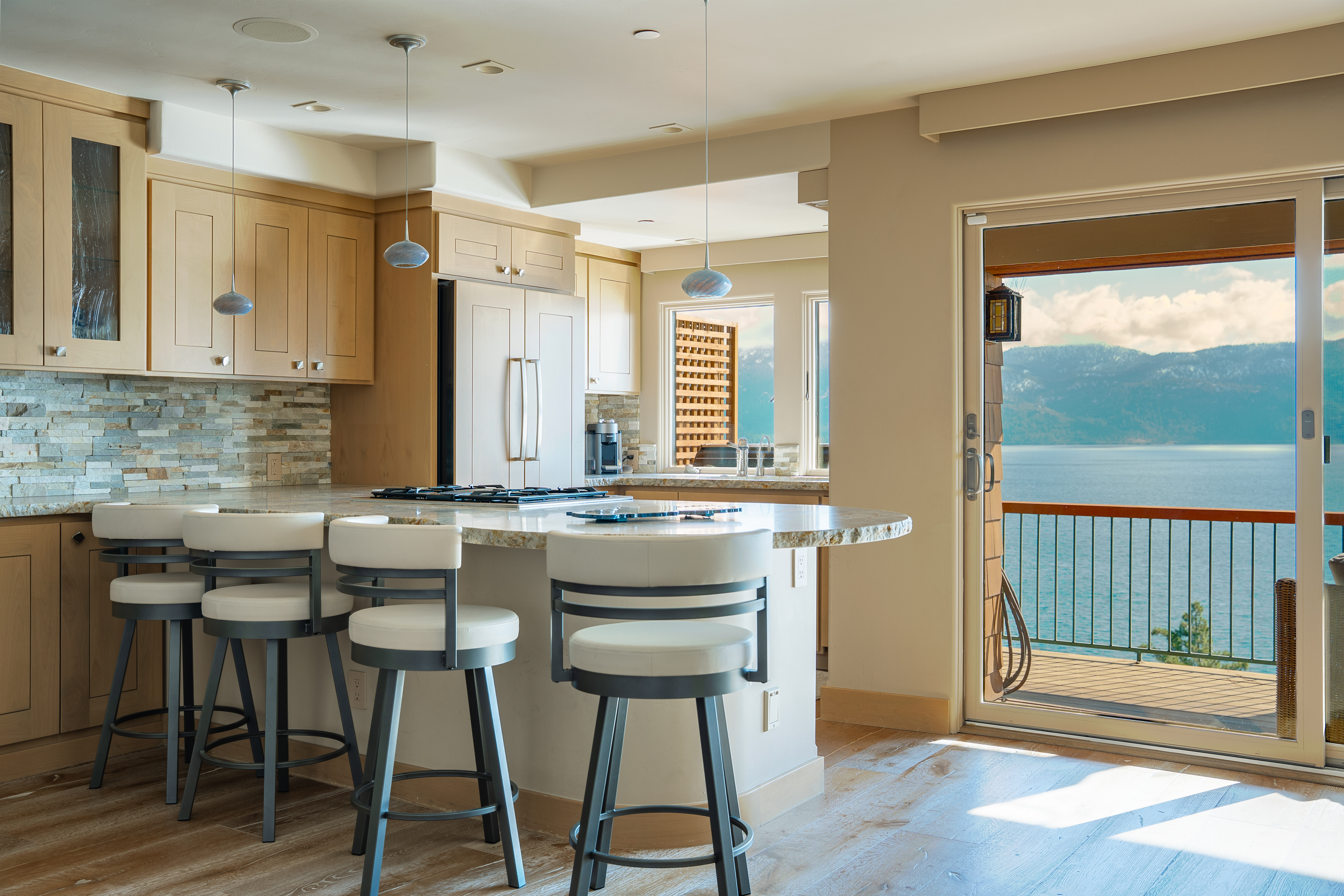 This is a bright and airy kitchen featuring light wood cabinetry, stone countertops, and a breakfast bar with modern stools. The kitchen opens to a balcony with stunning views of a lake and mountains, creating a seamless indoor-outdoor living experience. Stainless steel appliances and pendant lighting add a touch of modern elegance.