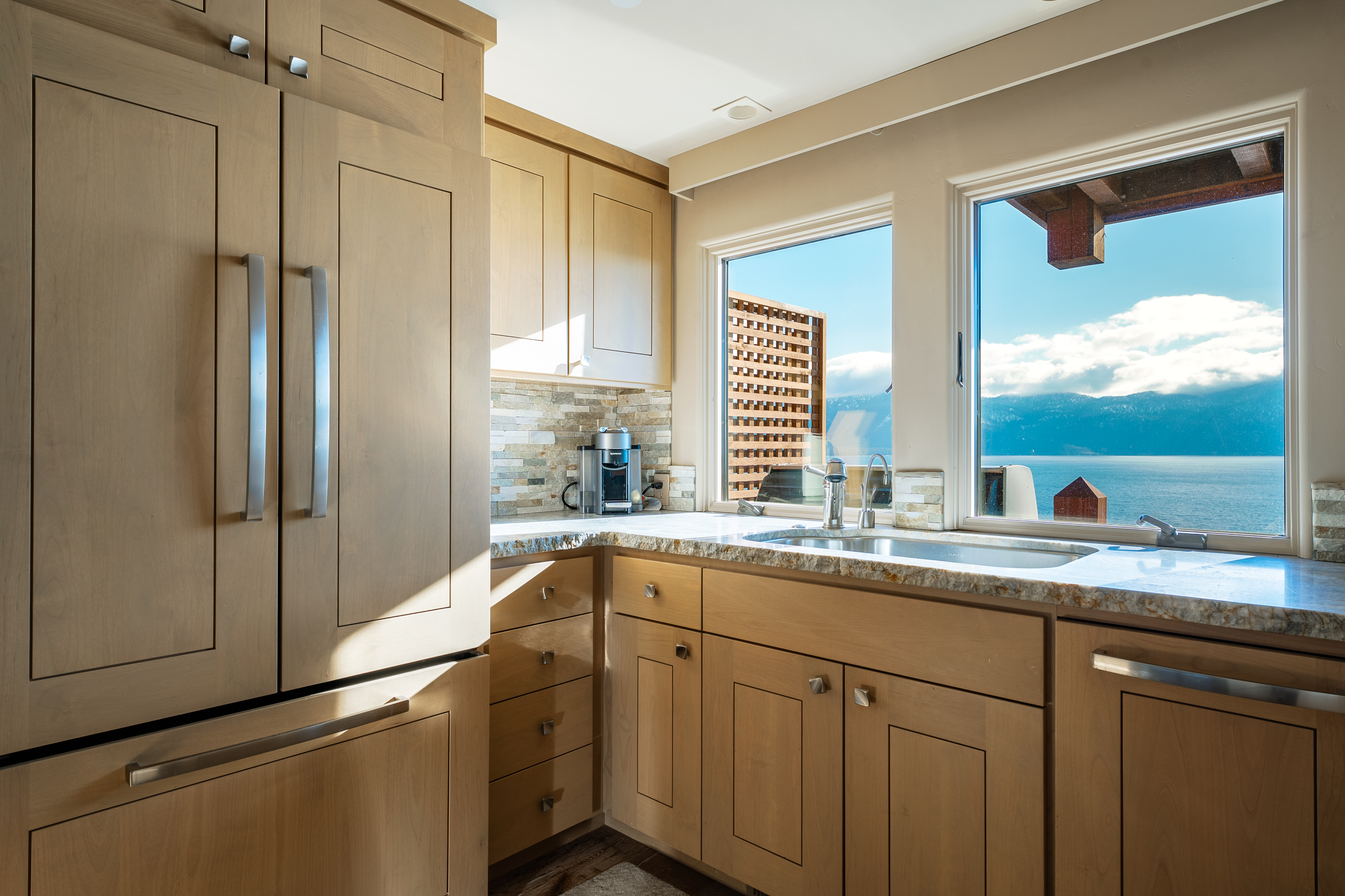 This is a well-lit kitchen featuring light wood cabinetry and granite countertops. A stainless steel refrigerator is on the left, and a sink is positioned in front of a window with a view of a lake and mountains. The kitchen has a clean and modern aesthetic.