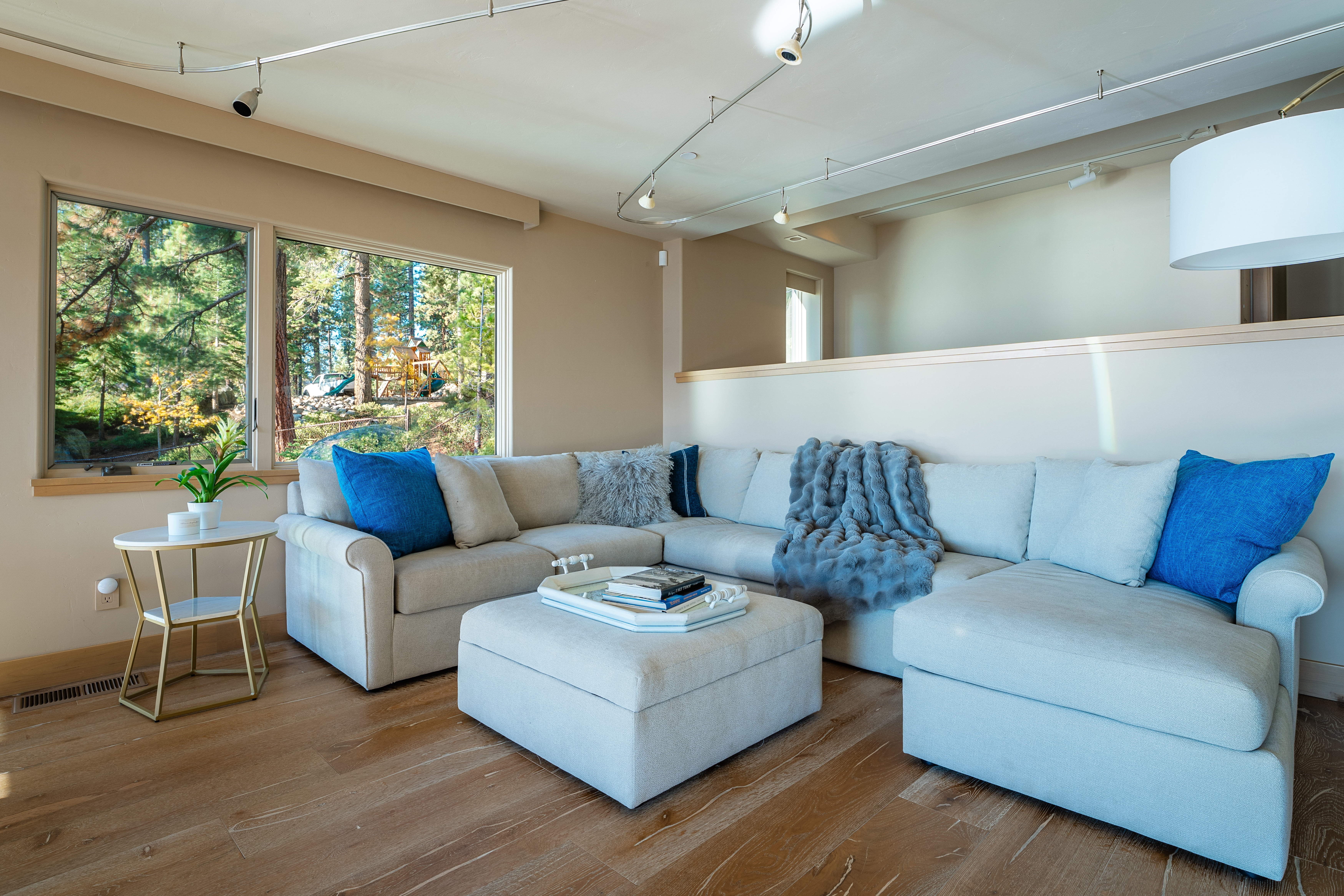 This is an interior shot of a living room featuring a large, sectional sofa with blue accent pillows and a gray faux fur throw. An ottoman sits in front of the sofa, topped with books and a tray. The room has hardwood floors and a large window offering a view of the outdoors, bringing natural light into the space.