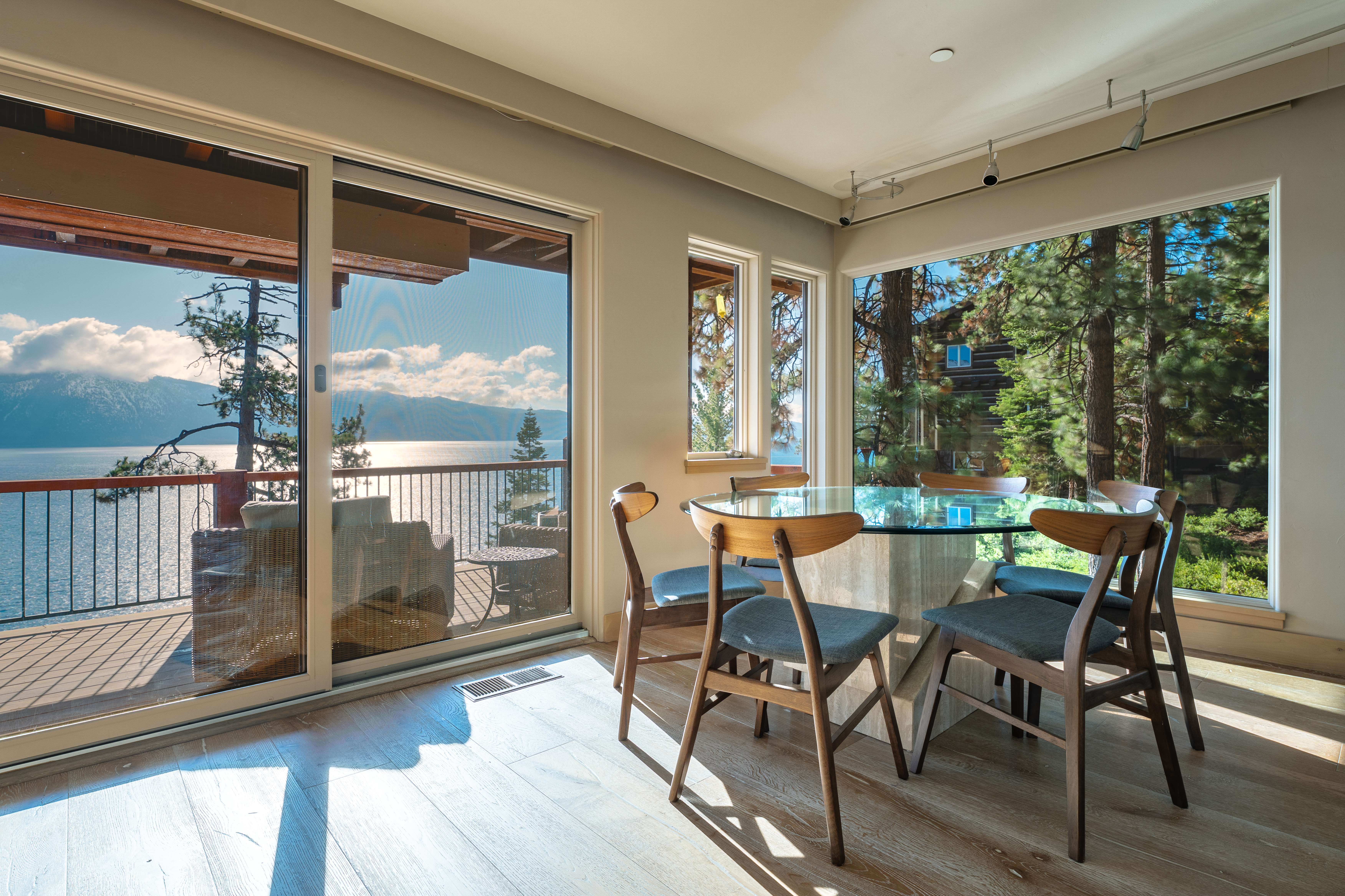 This dining room features a round glass table with a unique stone base, surrounded by modern wooden chairs with gray cushions. Large windows offer stunning views of the lake and surrounding trees, creating a bright and inviting atmosphere. Sliding glass doors lead to an outdoor deck, extending the living space.