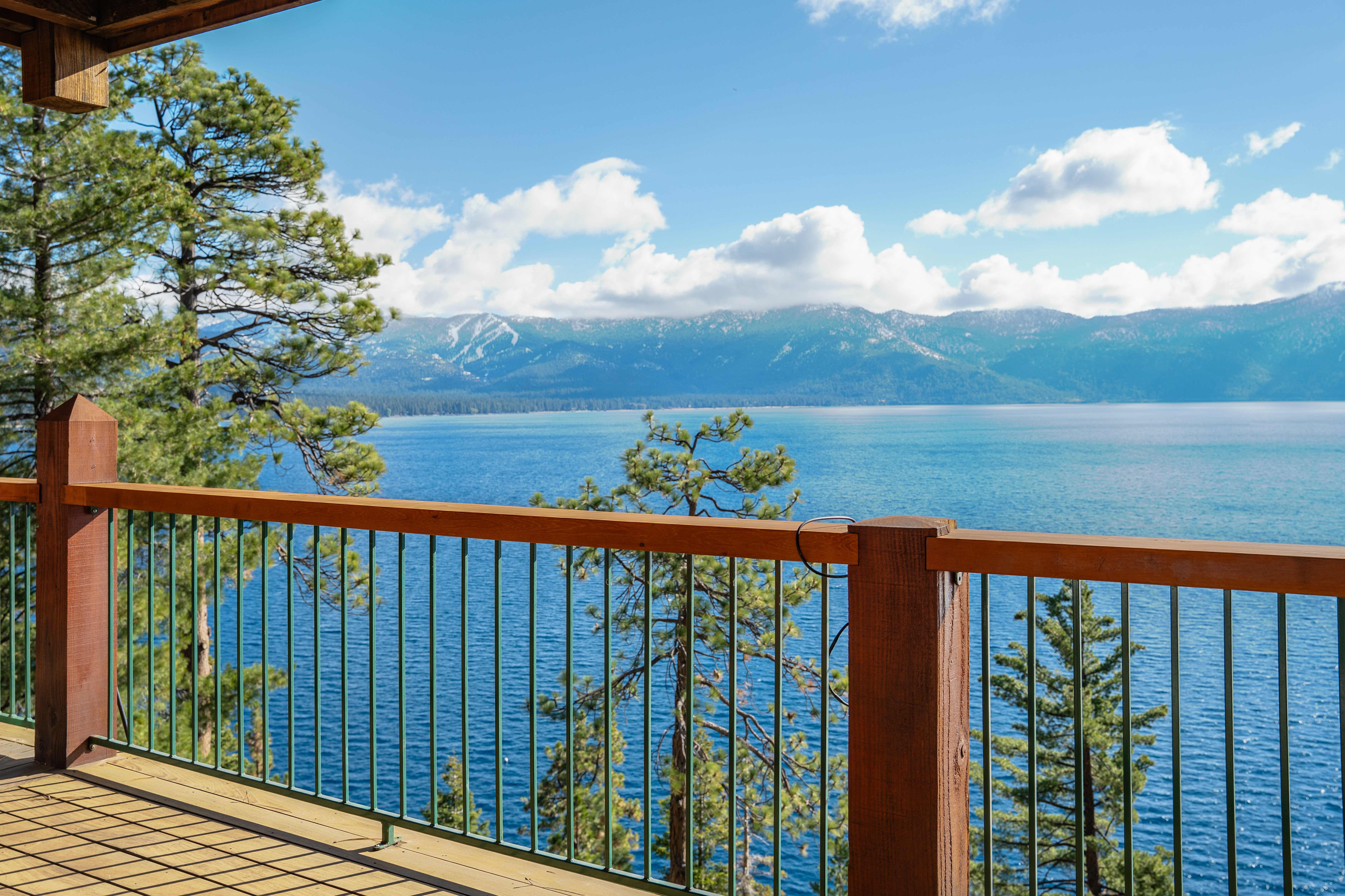 This image showcases a beautiful balcony view, overlooking a serene lake and distant mountains. The wooden railing and deck create a warm, inviting space, perfect for enjoying the scenic surroundings. The clear blue sky and calm water enhance the overall sense of tranquility and natural beauty.