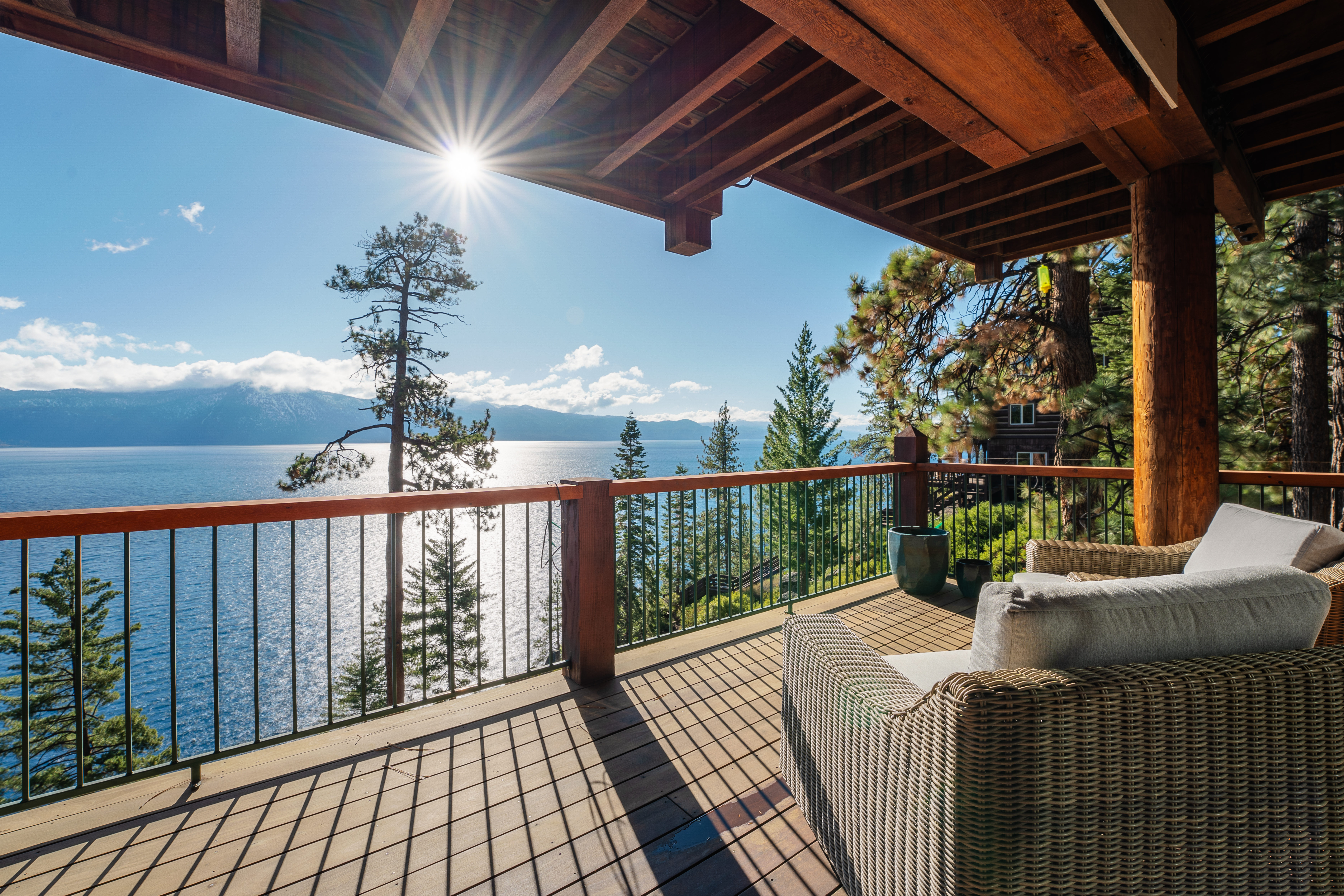 This image showcases a stunning outdoor deck with breathtaking lake views. The deck features a wooden floor, a dark metal railing, and comfortable wicker furniture with white cushions. The sun shines brightly over the lake, creating a serene and inviting atmosphere, perfect for relaxation and enjoying the natural surroundings.