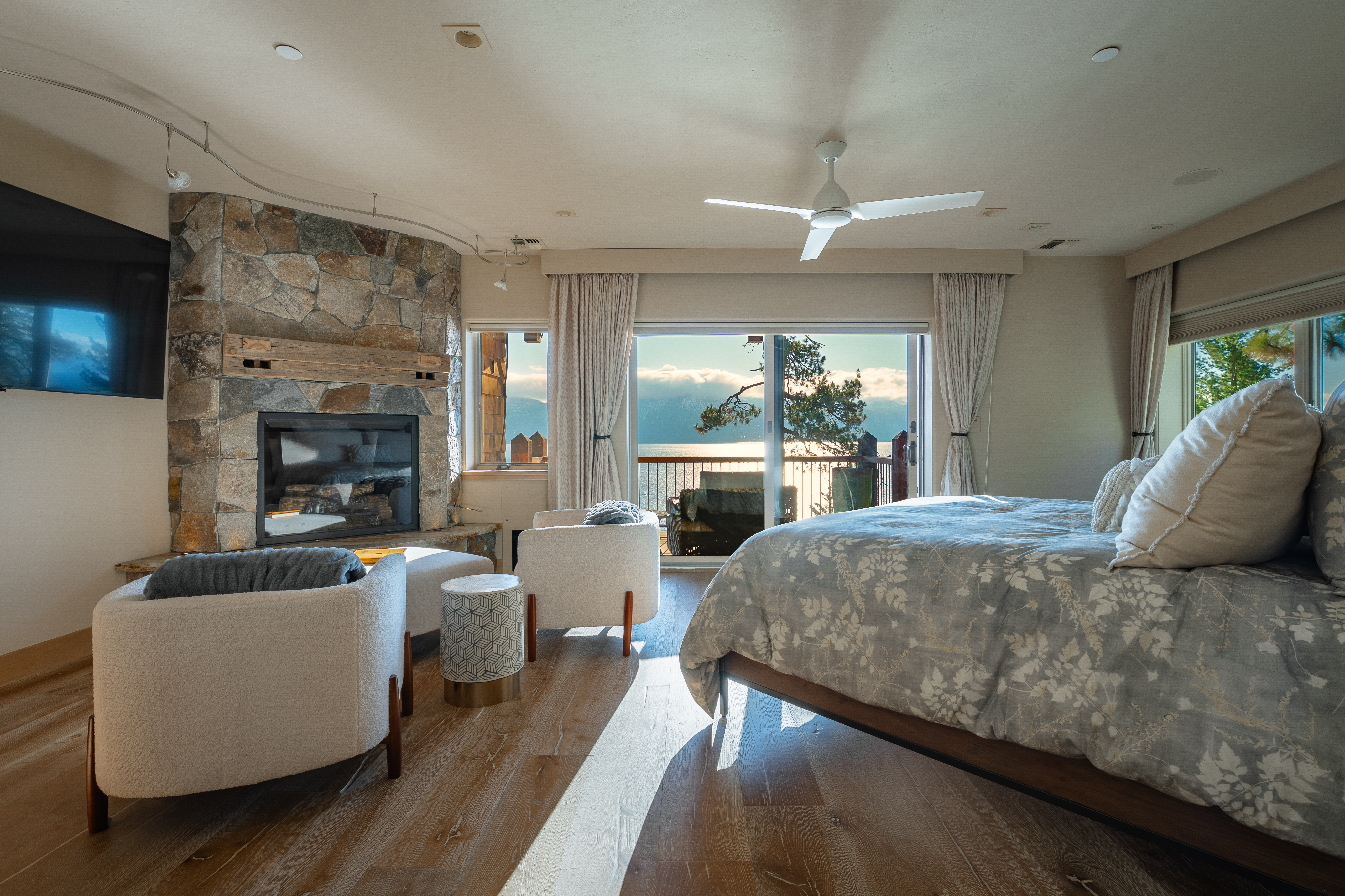 This is a primary bedroom featuring a stone fireplace, two armchairs, and a bed with a floral patterned duvet. Large windows offer a view of a lake and mountains. The room has hardwood floors and a ceiling fan, creating a cozy and luxurious atmosphere.
