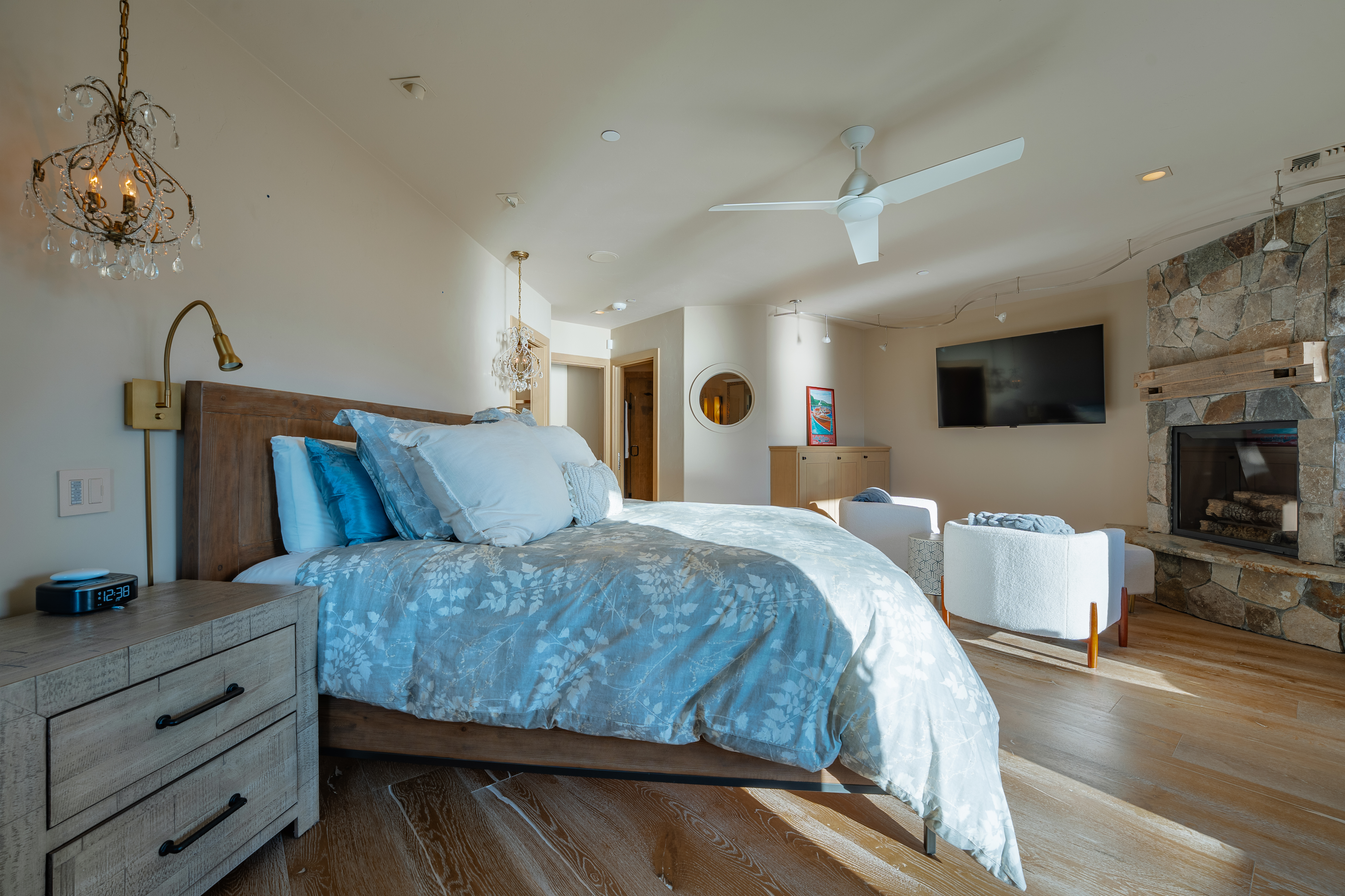 This is a primary bedroom featuring a large bed with blue and white floral bedding, a wooden headboard, and a nightstand with a digital clock. The room includes a stone fireplace, two white armchairs, and a ceiling fan, creating a cozy and luxurious atmosphere. The flooring is light wood, and the room is well-lit with natural light and decorative chandeliers.