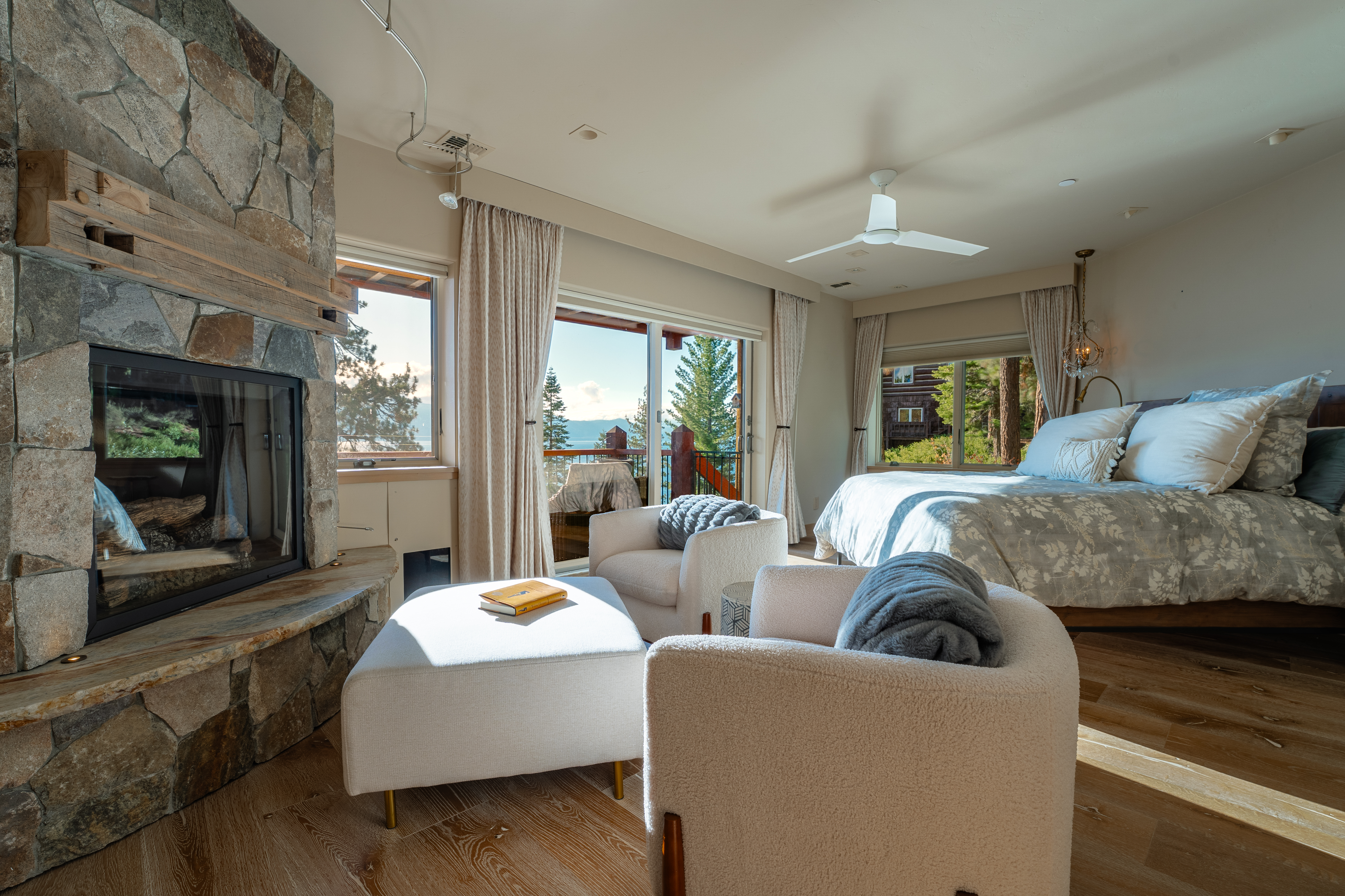 This is a bright and airy primary bedroom featuring a stone fireplace, a king-size bed with a floral duvet, and two cozy armchairs with a matching ottoman. Large windows and a sliding glass door offer views of the surrounding landscape and a balcony, enhancing the room's serene and luxurious atmosphere. The hardwood floors and neutral color palette contribute to the room's inviting and sophisticated style.