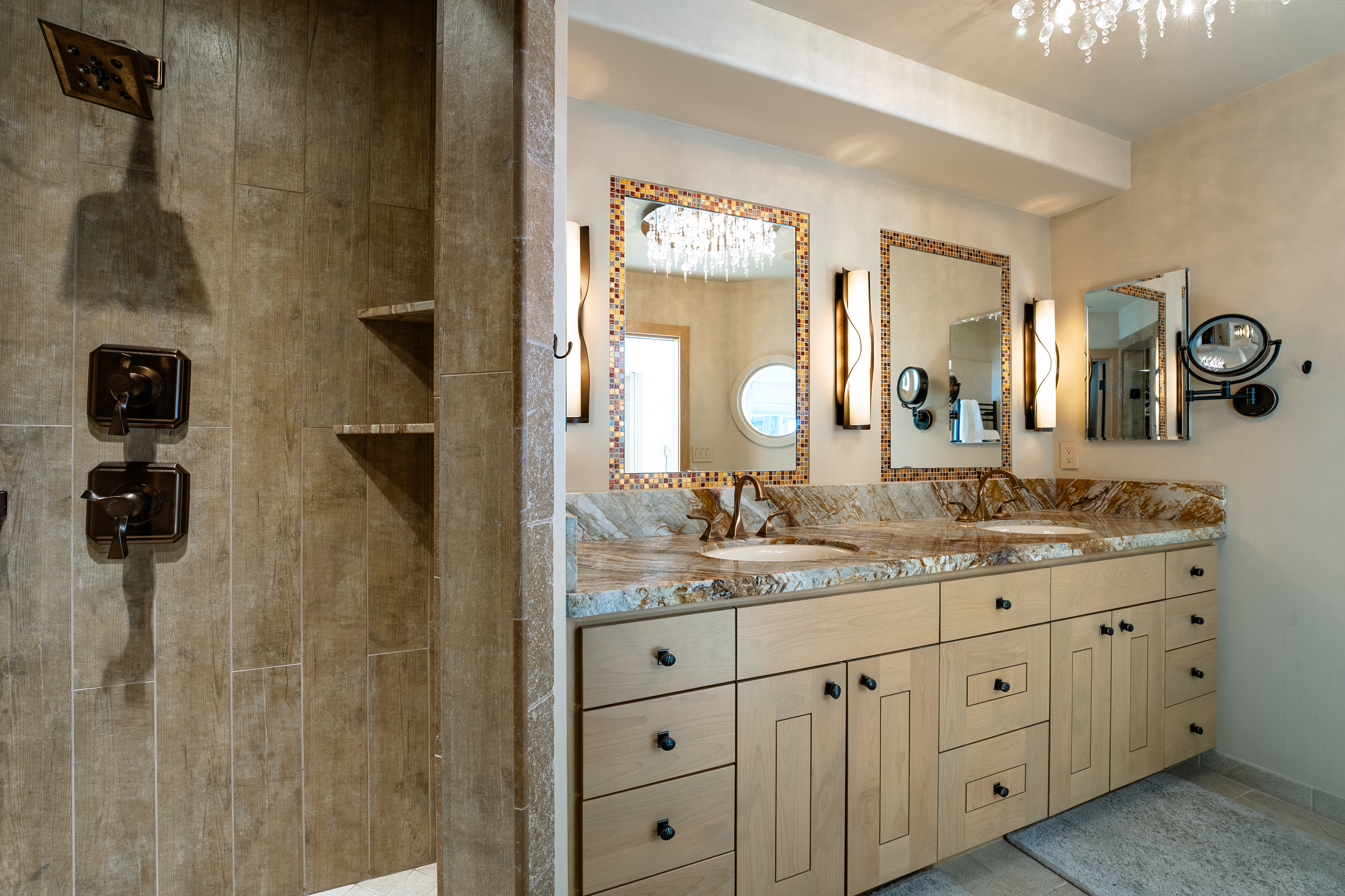 This is a well-appointed primary bathroom featuring a double vanity with a granite countertop and light wood cabinetry. The mirrors are framed with decorative mosaic tiles, and sconces provide warm lighting. A walk-in shower with bronze fixtures and built-in shelves is visible to the left, creating a luxurious and functional space.