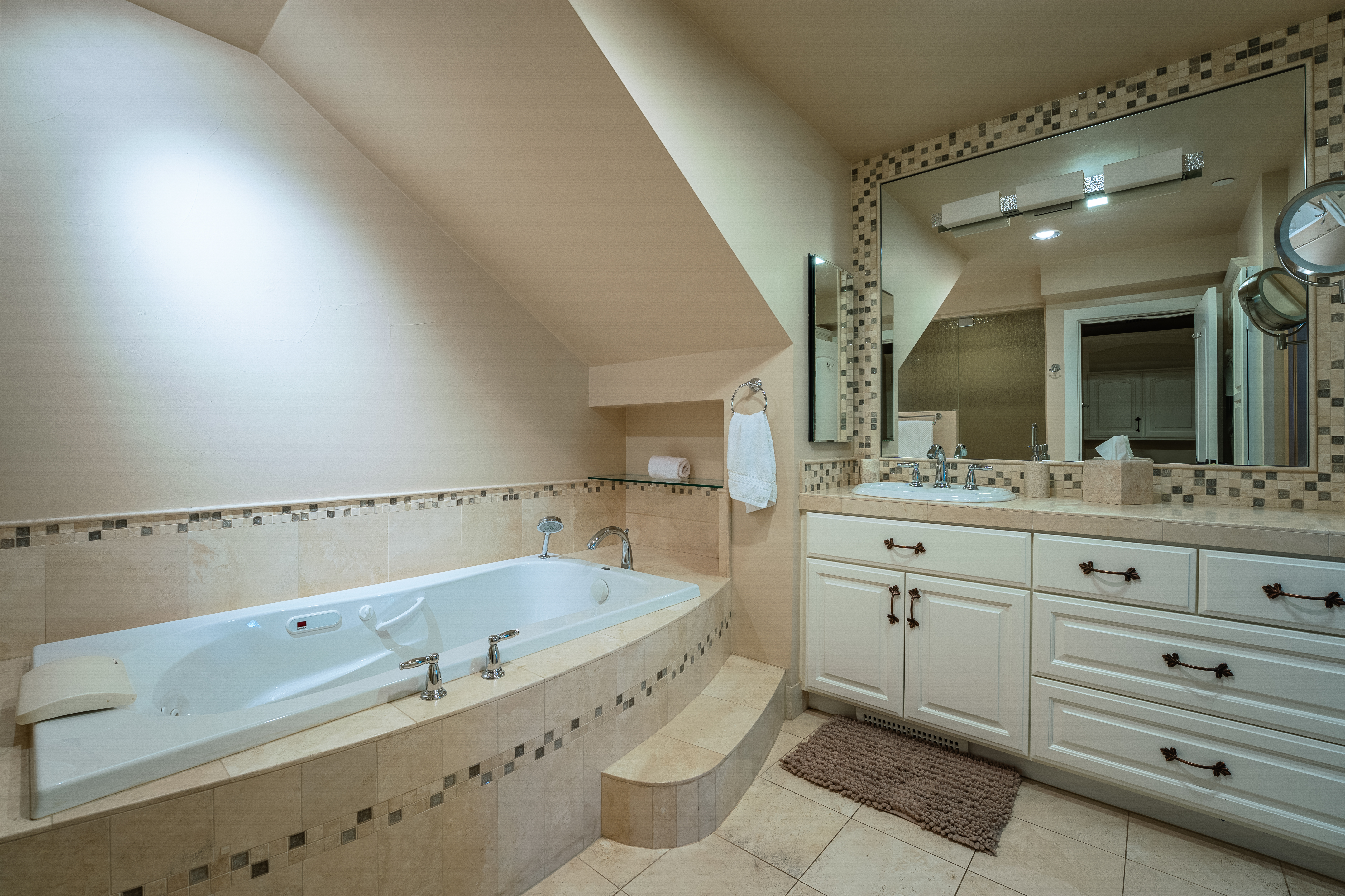 This is a well-lit primary bathroom featuring a large soaking tub with tile surround and steps for easy access. A vanity with ample storage, a sink, and a large mirror with decorative tile border are also present. The room has a neutral color palette, creating a relaxing and spa-like atmosphere.
