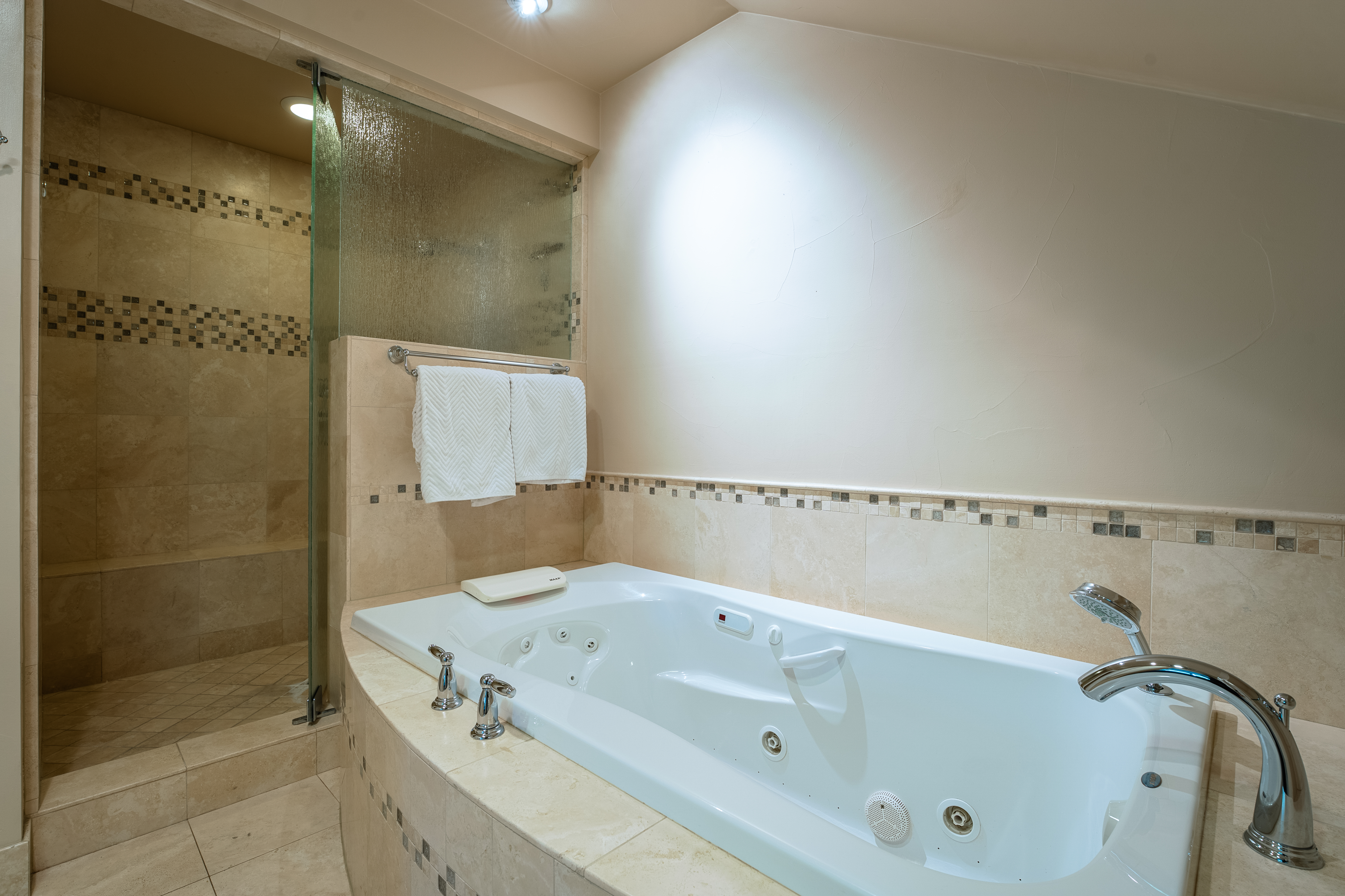 This primary bathroom features a large, white jacuzzi tub with chrome fixtures, surrounded by beige tile with a decorative mosaic border. A glass-enclosed shower is visible to the left, and two white towels hang on a towel rack above the tub. The overall impression is clean and luxurious.