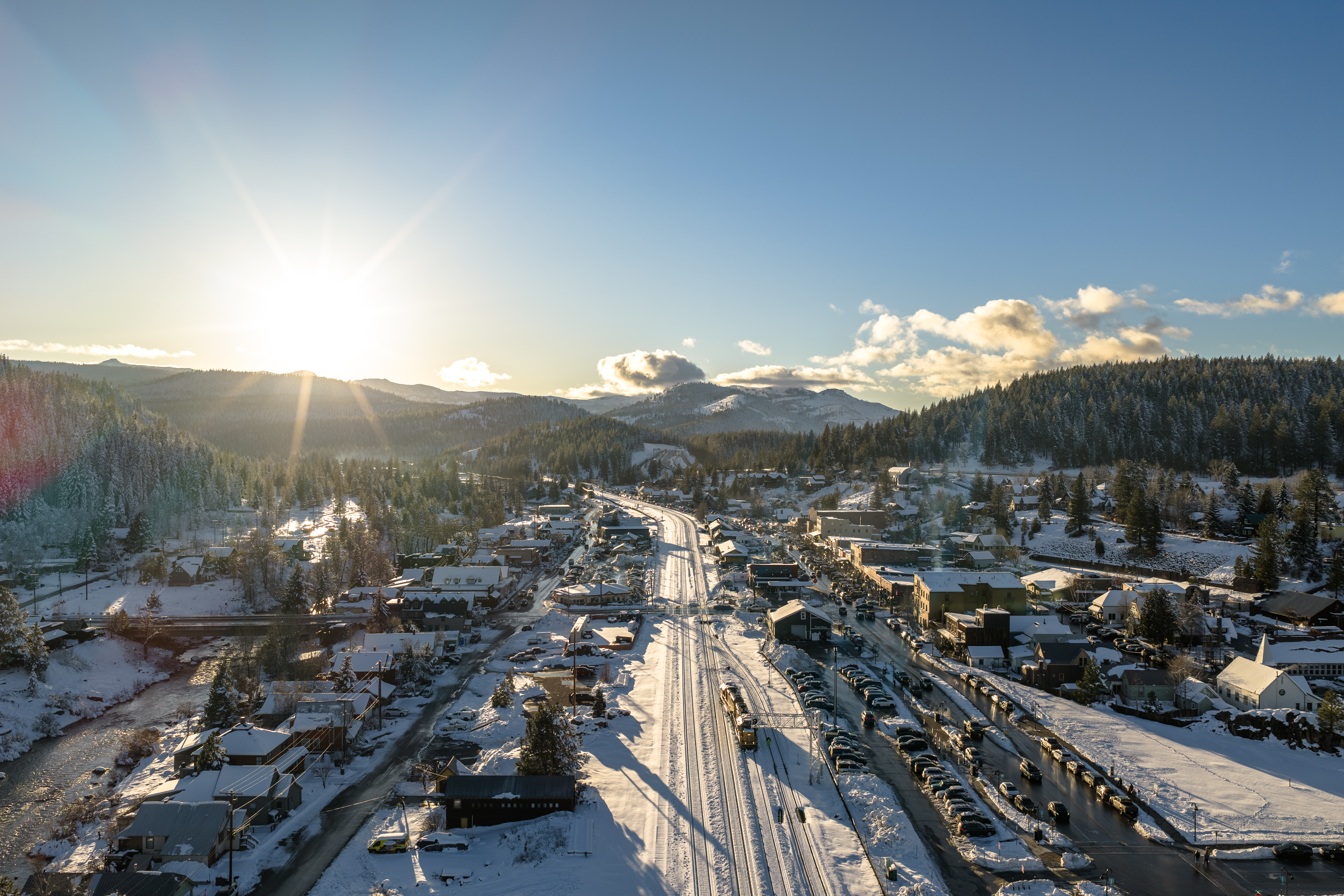 This aerial view showcases a charming town nestled amidst snow-covered mountains and evergreen forests. The main street is lined with buildings, and a railway runs through the center, adding to the town's character. A large parking lot filled with cars suggests a bustling community, while the bright sunlight creates a warm and inviting atmosphere.