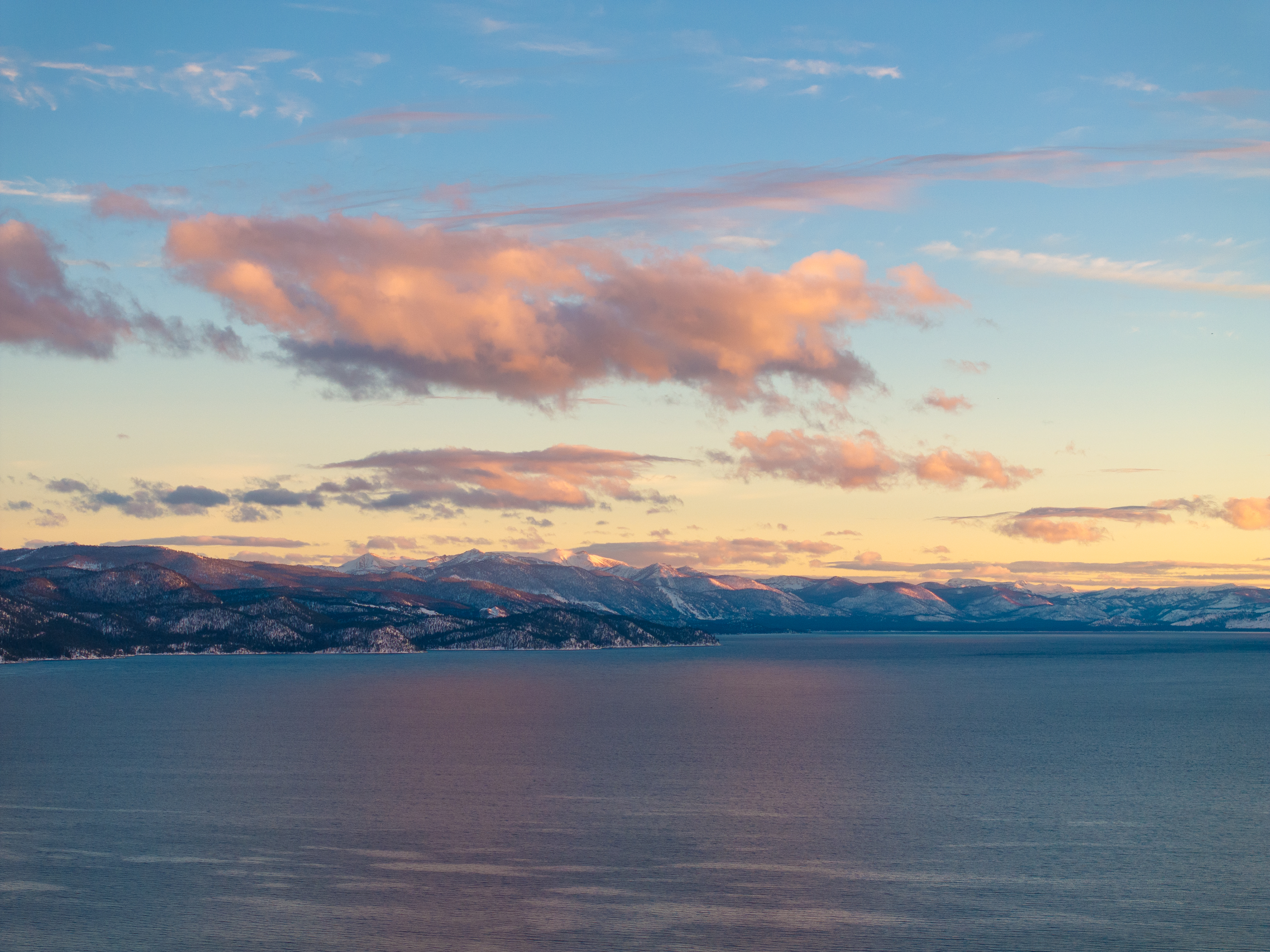 This aerial view showcases a serene lake surrounded by snow-capped mountains under a pastel-colored sky at sunset. The calm water reflects the soft light, creating a tranquil and picturesque scene. The landscape suggests a peaceful and secluded location, ideal for a waterfront property.