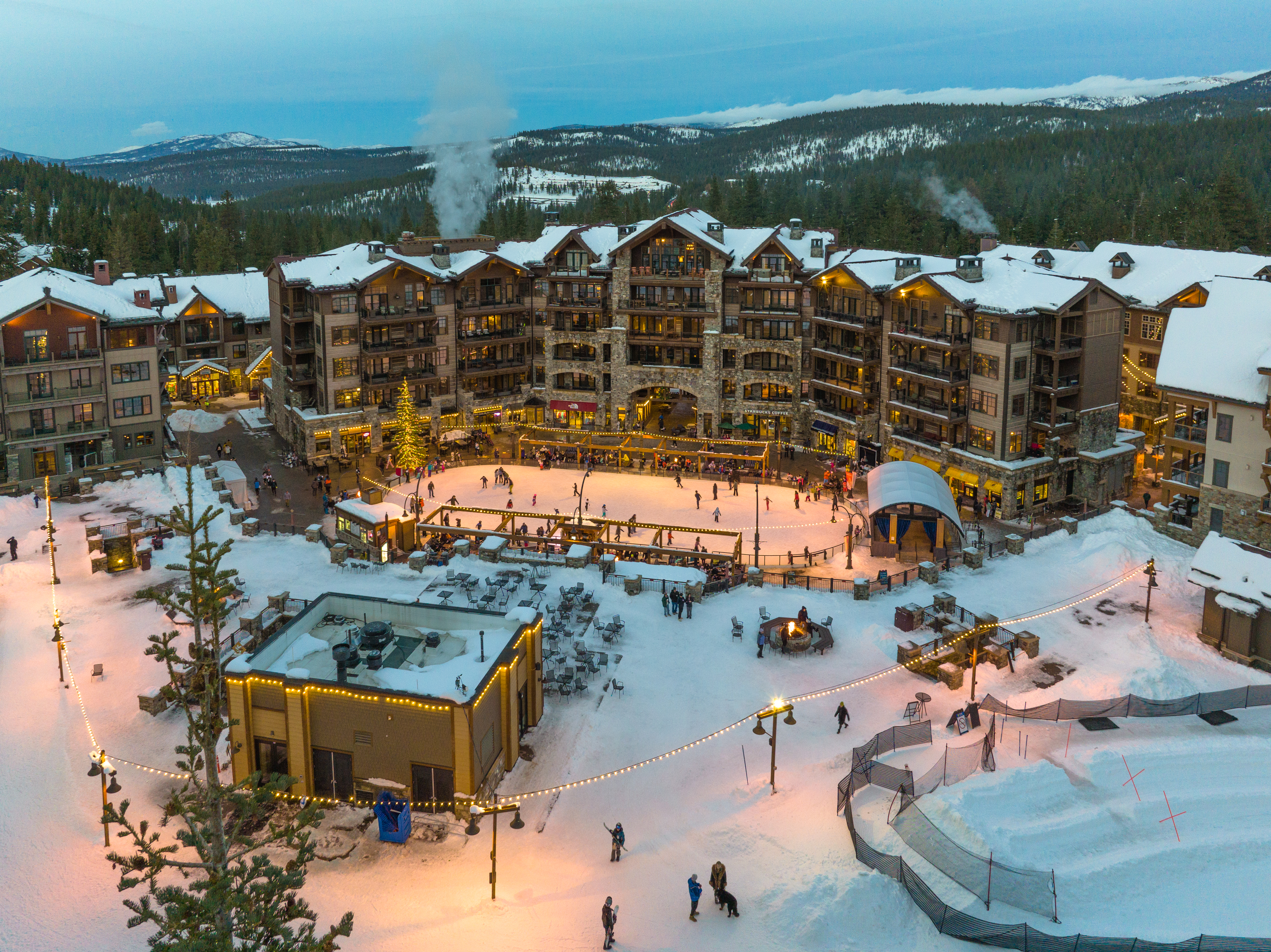 This aerial view showcases a luxurious ski resort village at dusk, complete with snow-covered buildings, an ice skating rink, and festive lighting. The multi-story buildings feature stone and wood accents, creating a rustic yet elegant aesthetic. The scene is lively with people enjoying the winter activities, and the surrounding snow-covered mountains add to the picturesque setting.