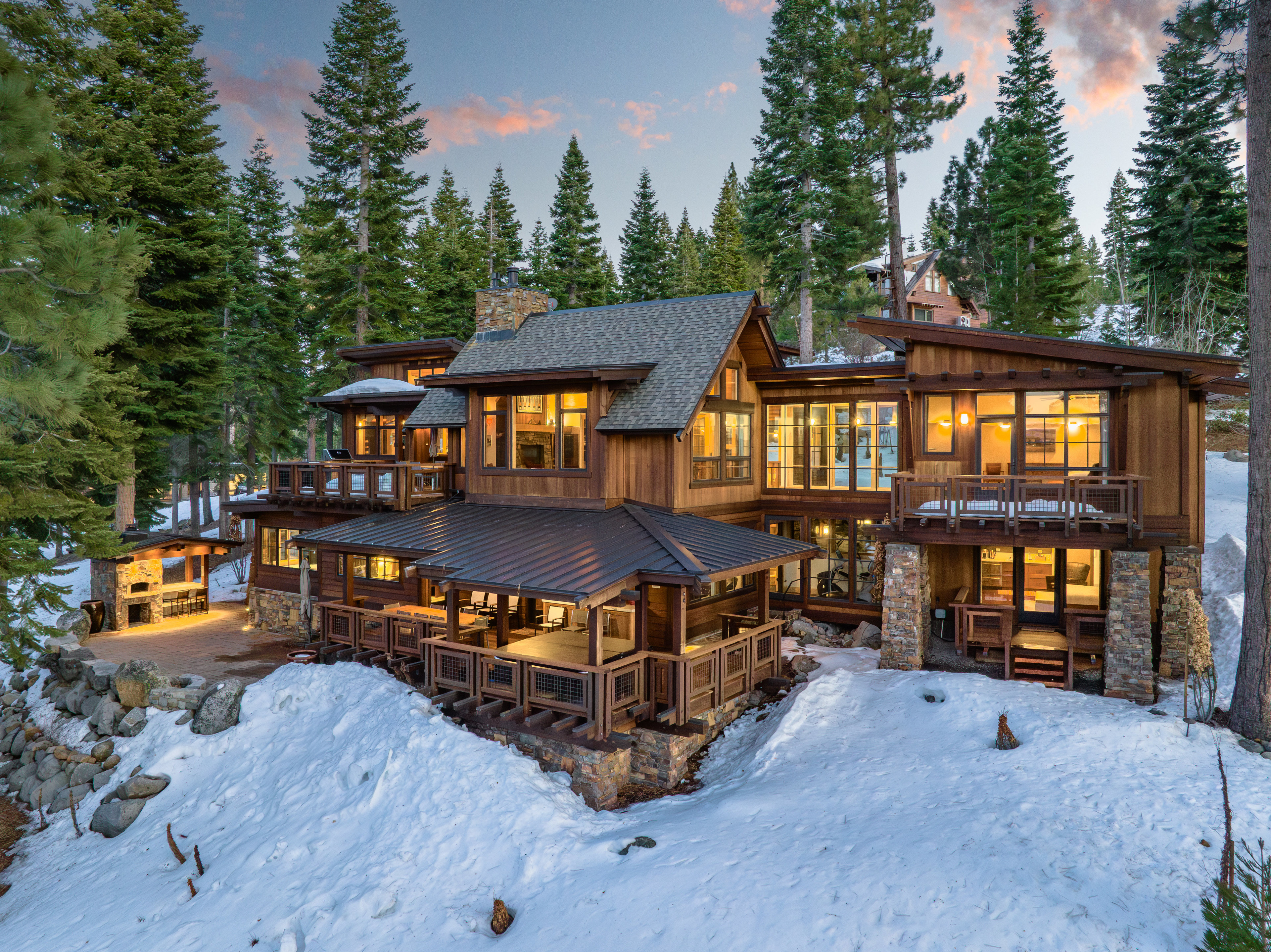 This is a rear exterior view of a luxurious multi-level home nestled among tall evergreen trees with snow on the ground. The house features wood siding, stone accents, multiple decks and balconies, and large windows that offer stunning views. The architecture is mountain modern, blending seamlessly with the natural surroundings.