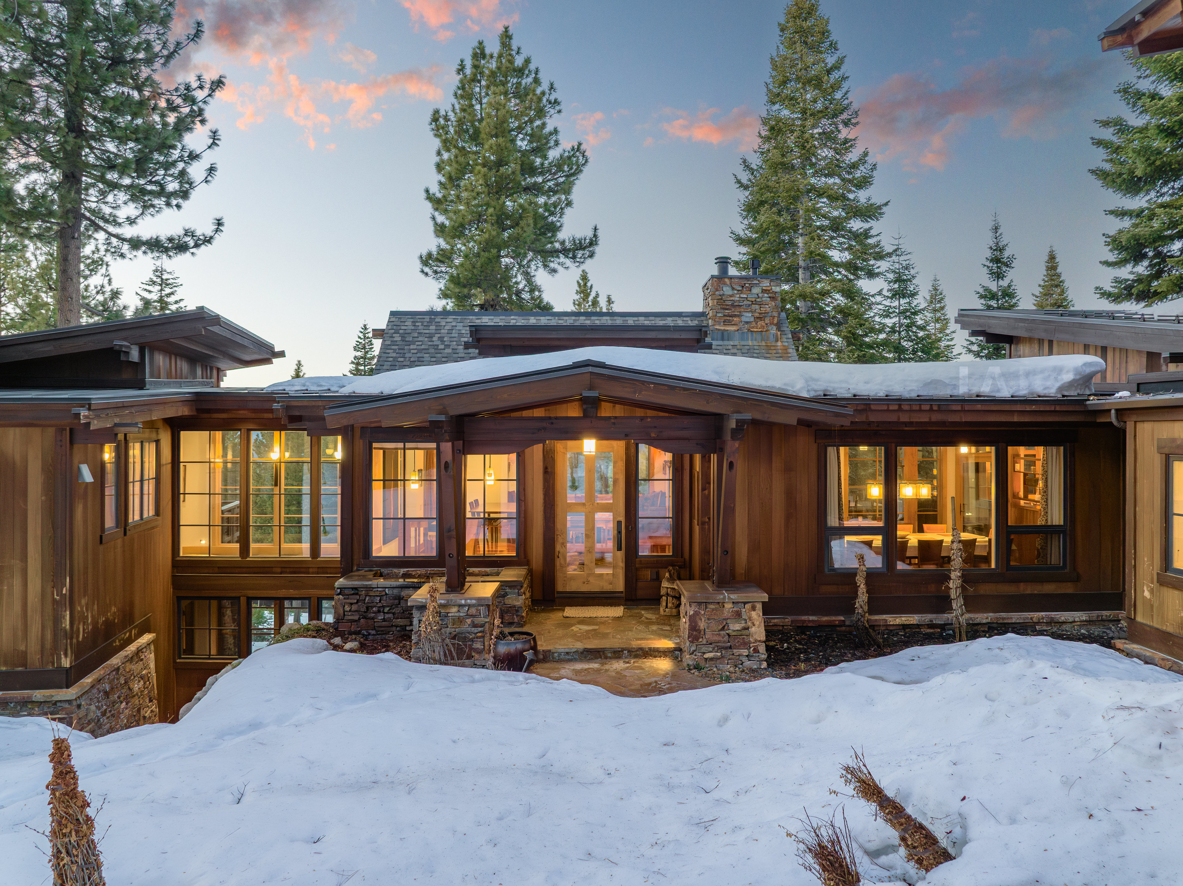 The image showcases the front exterior of a luxurious mountain home during winter. The house features a wood facade, stone accents, and large windows that allow natural light to flood the interior. Snow covers the ground, adding to the serene and picturesque setting, while the architecture blends seamlessly with the surrounding natural landscape.