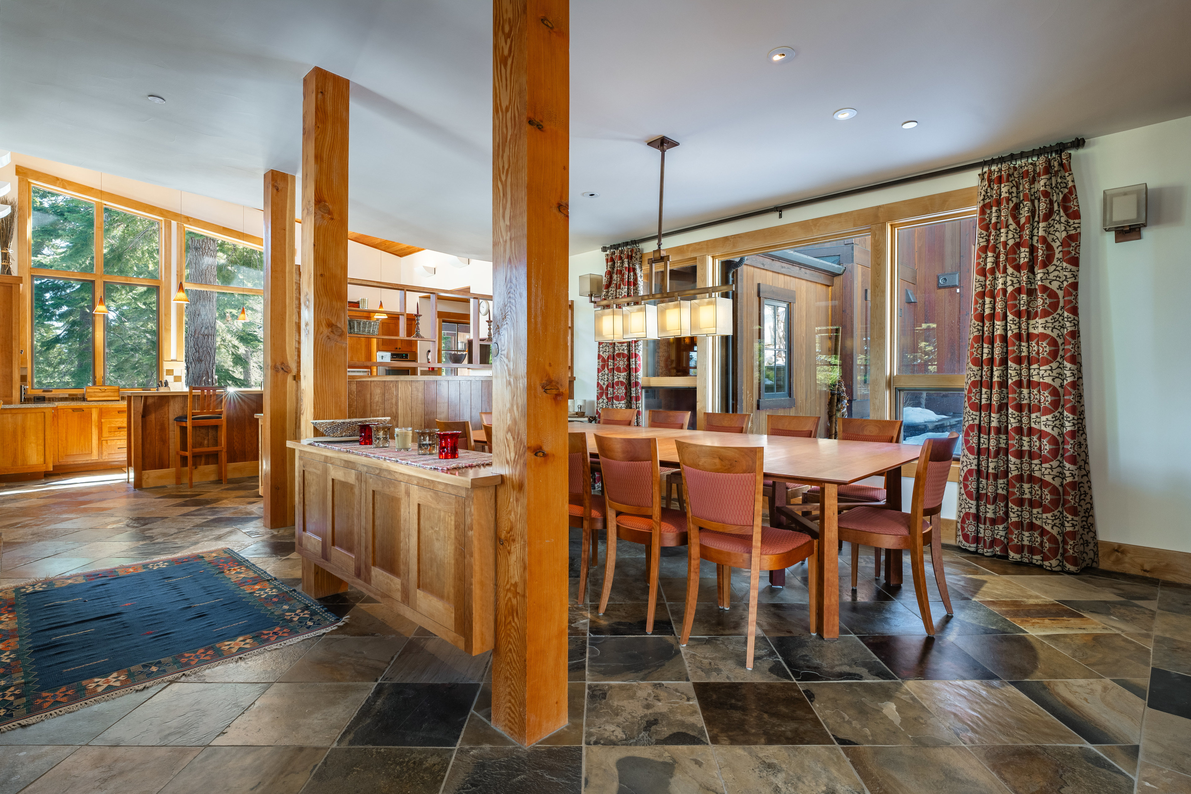 This interior shot showcases a dining room with a rustic yet elegant design. The room features a large wooden dining table surrounded by chairs with patterned cushions, complemented by a unique light fixture overhead. Natural light streams in through the windows, highlighting the stone tile flooring and wooden support beams, creating a warm and inviting atmosphere.