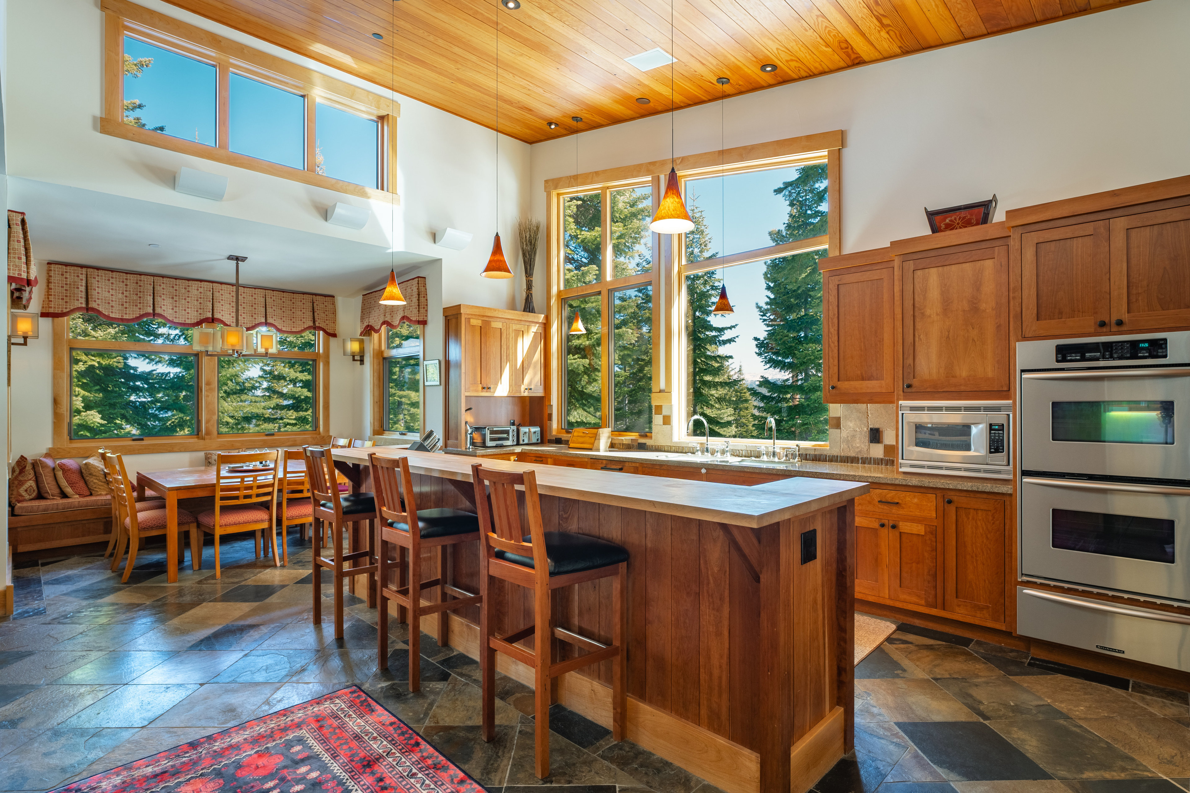 This is a well-lit kitchen featuring wooden cabinetry, stainless steel appliances, and a large island with bar seating. Large windows offer a view of the surrounding trees, and pendant lights hang above the island. The floor is tiled with a dark stone, and the overall style is warm and inviting.