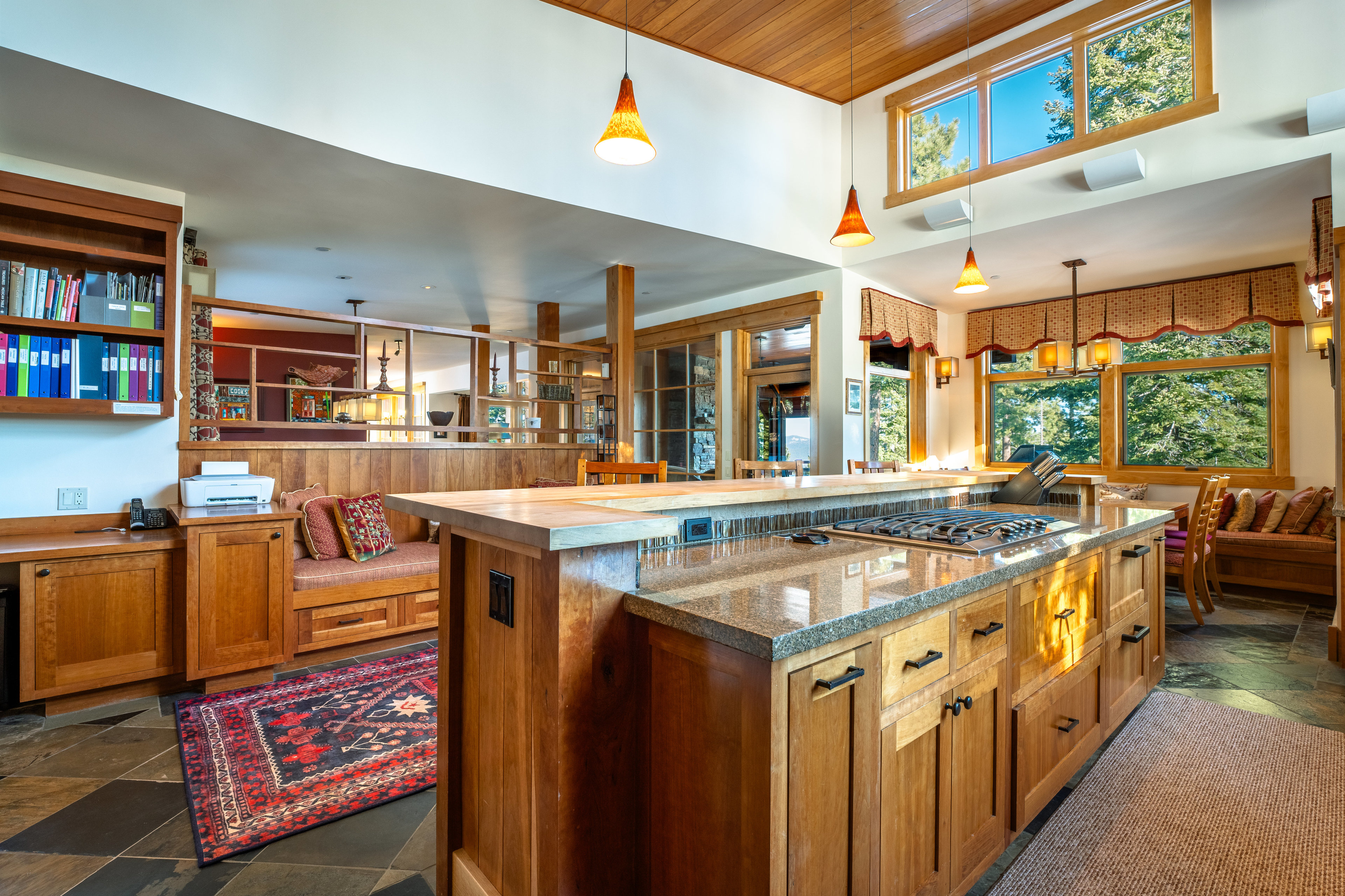 This interior shot showcases a spacious kitchen with warm wood cabinetry, granite countertops, and a central island featuring a cooktop. The kitchen seamlessly blends into an adjacent dining area, highlighted by large windows offering natural light and views of the outdoors. The overall design emphasizes a comfortable and inviting atmosphere, perfect for both cooking and entertaining.
