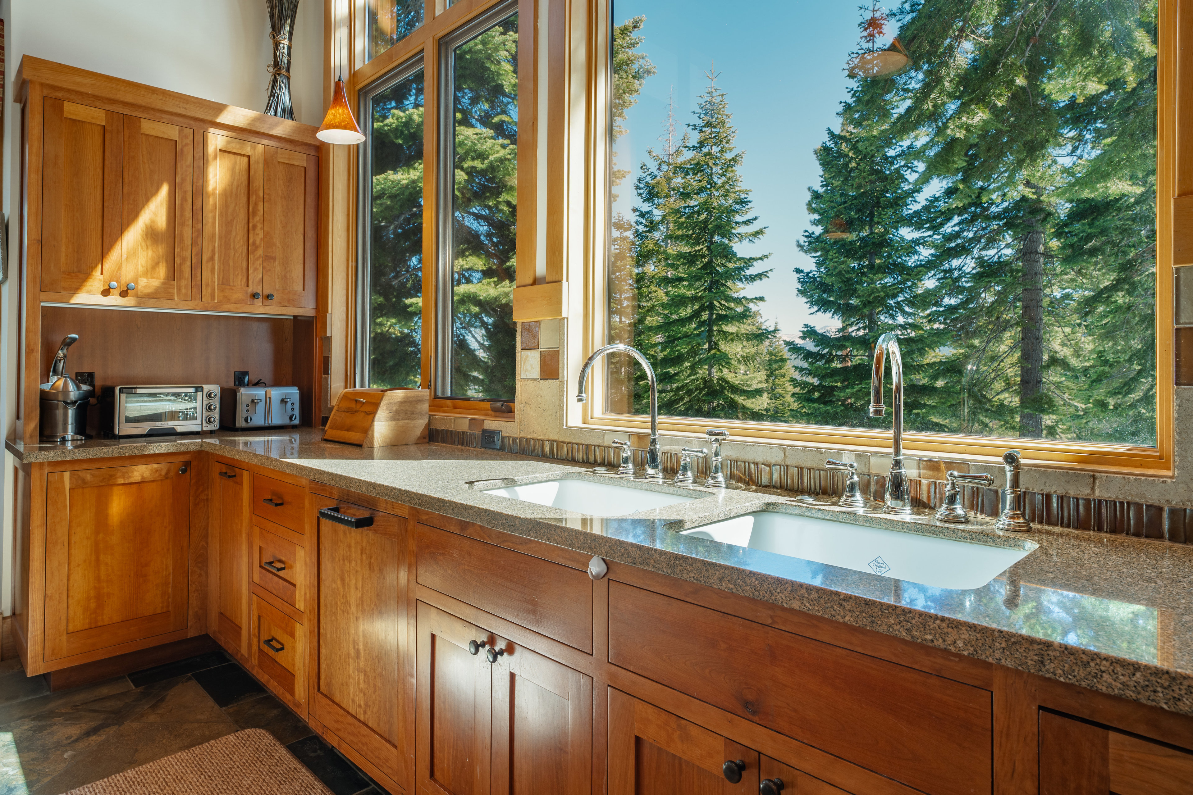 This is a well-lit kitchen featuring wooden cabinetry and granite countertops. A large window provides a view of evergreen trees, bringing natural light into the space. The kitchen is equipped with stainless steel appliances and a double sink, creating a warm and inviting atmosphere.