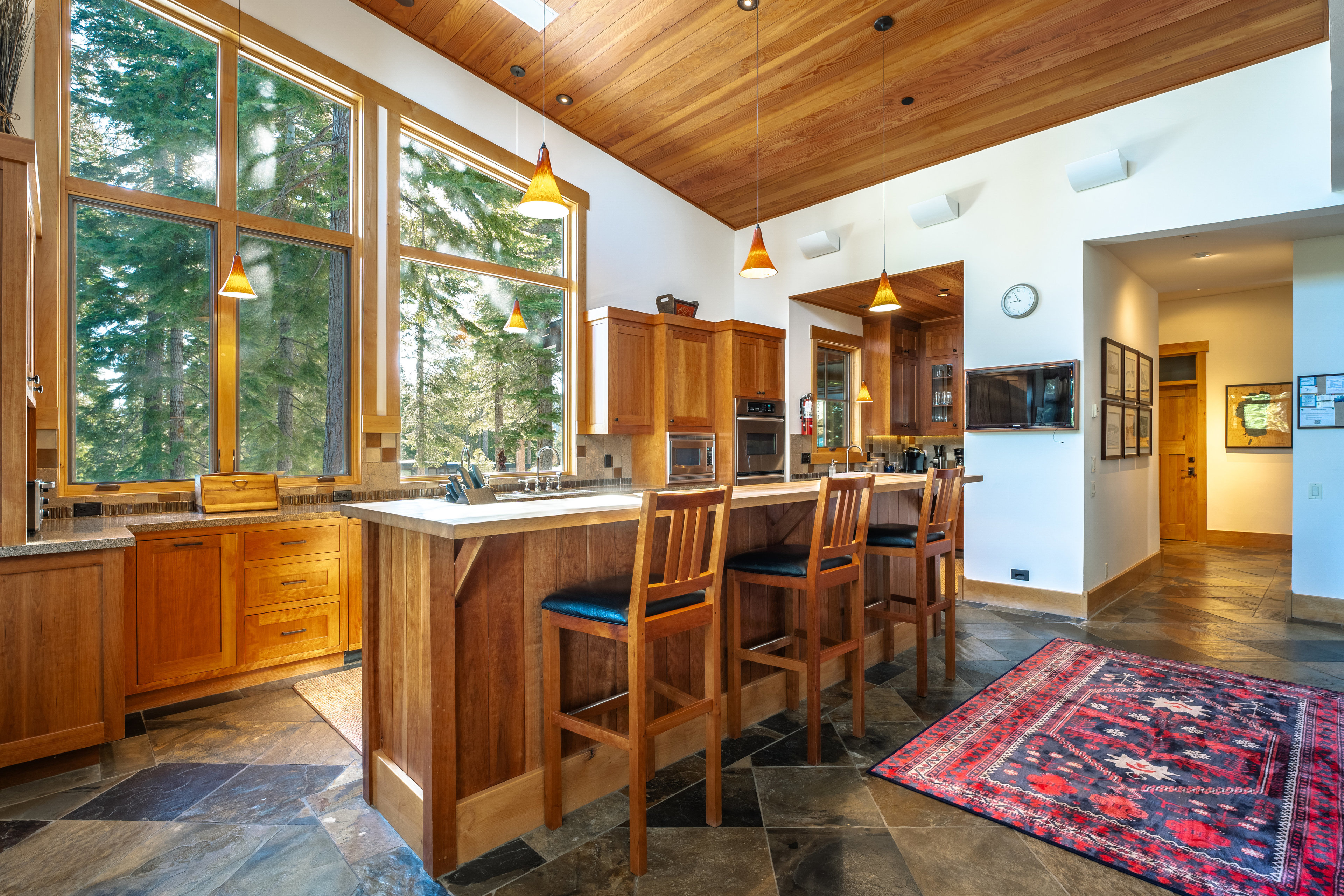 This is a warm and inviting kitchen featuring wood cabinetry and a large island with bar seating. The kitchen is well-lit with natural light from large windows overlooking a wooded area, complemented by pendant lighting. The flooring is a unique tile pattern, and a decorative rug adds a touch of color to the space.