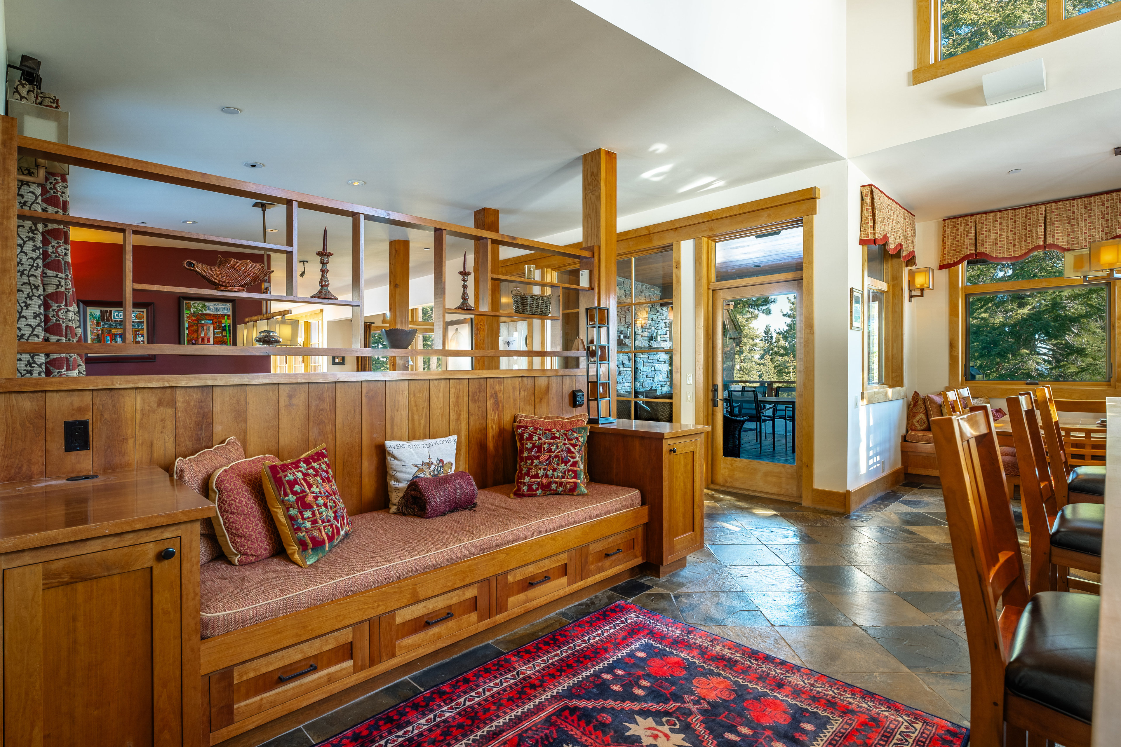 This interior shot showcases a cozy living room area with a built-in wooden bench seat featuring storage drawers and decorative pillows. A wooden partition with shelving separates the space, while a patterned rug adds warmth to the tiled floor. Natural light streams in through a nearby door, highlighting the room's inviting atmosphere.