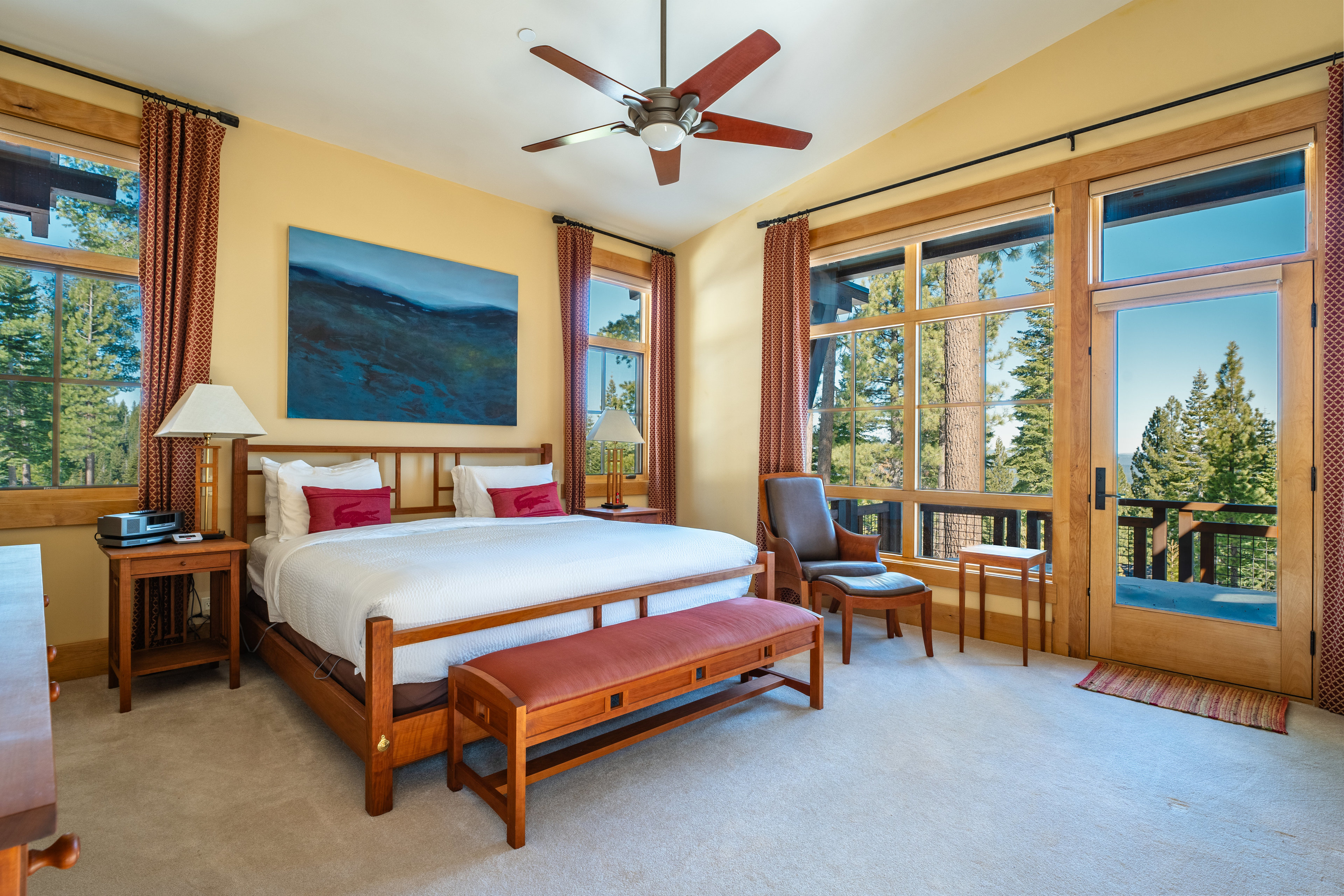 This is a primary bedroom featuring a wooden bed frame with white linens and red decorative pillows. An abstract painting hangs above the bed, and the room is illuminated by natural light streaming through large windows with wooden frames. The room also includes a wooden bench at the foot of the bed, a leather chair, and bedside tables with lamps, creating a warm and inviting atmosphere.