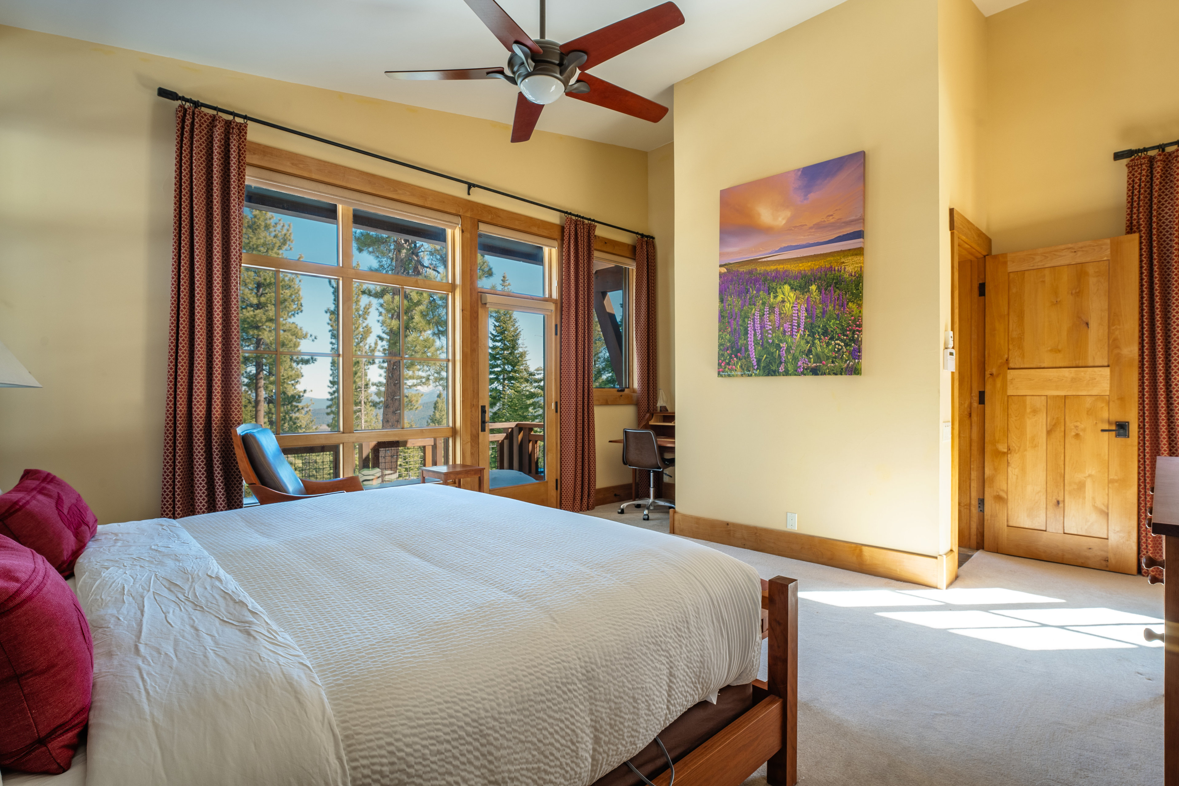 This is a primary bedroom featuring a large bed with white linens and red pillows, a ceiling fan, and a wall painting. The room has large windows offering a view of the outdoors, with wooden frames and red curtains. A wooden door and a desk area are also visible, contributing to the room's warm and inviting atmosphere.
