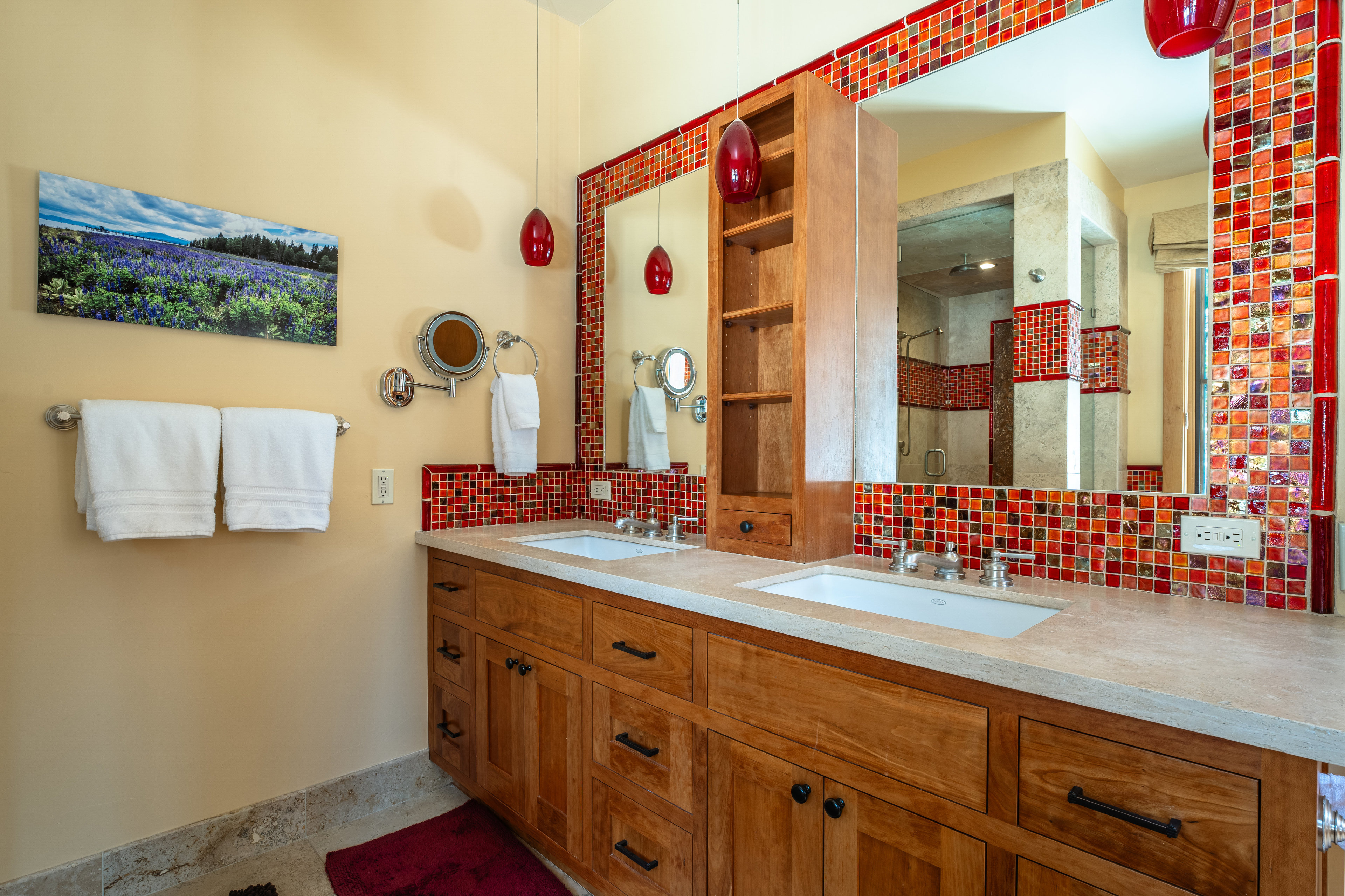 This is a primary bathroom featuring a double vanity with wooden cabinets and a light-colored countertop. The mirrors are framed with red and orange mosaic tiles, adding a vibrant touch. Two red pendant lights hang above the vanity, and a landscape painting adorns the wall, creating a serene atmosphere.