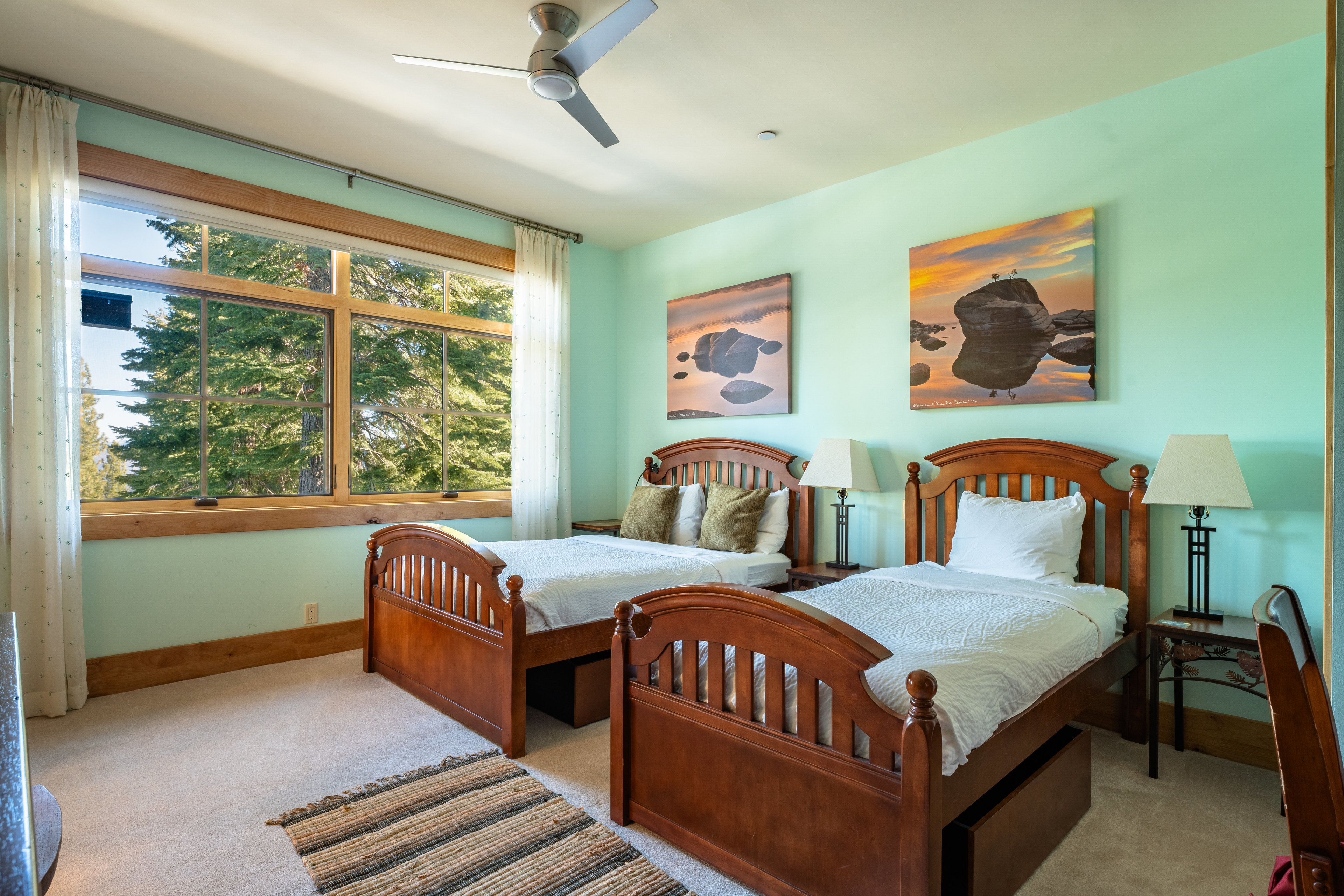 This is a cozy guest bedroom featuring two wooden twin beds with white bedding and decorative pillows. Artwork hangs above each bed, and a large window provides natural light and a view of the trees outside. The walls are painted a calming light green, and a ceiling fan adds to the comfort of the room.
