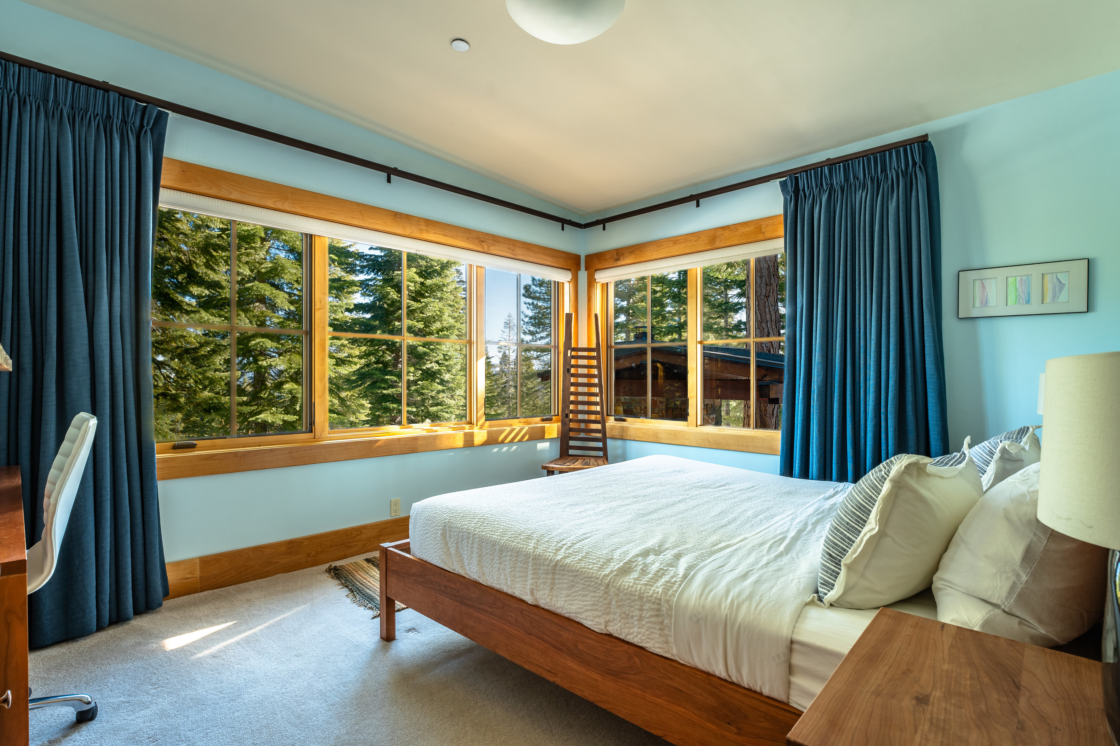 This is a bright and airy primary bedroom featuring a large bed with white linens, wood frame, and several pillows. Large windows with wood frames offer views of the surrounding trees, and blue curtains frame the windows. The room is painted a light blue, creating a serene and inviting atmosphere.