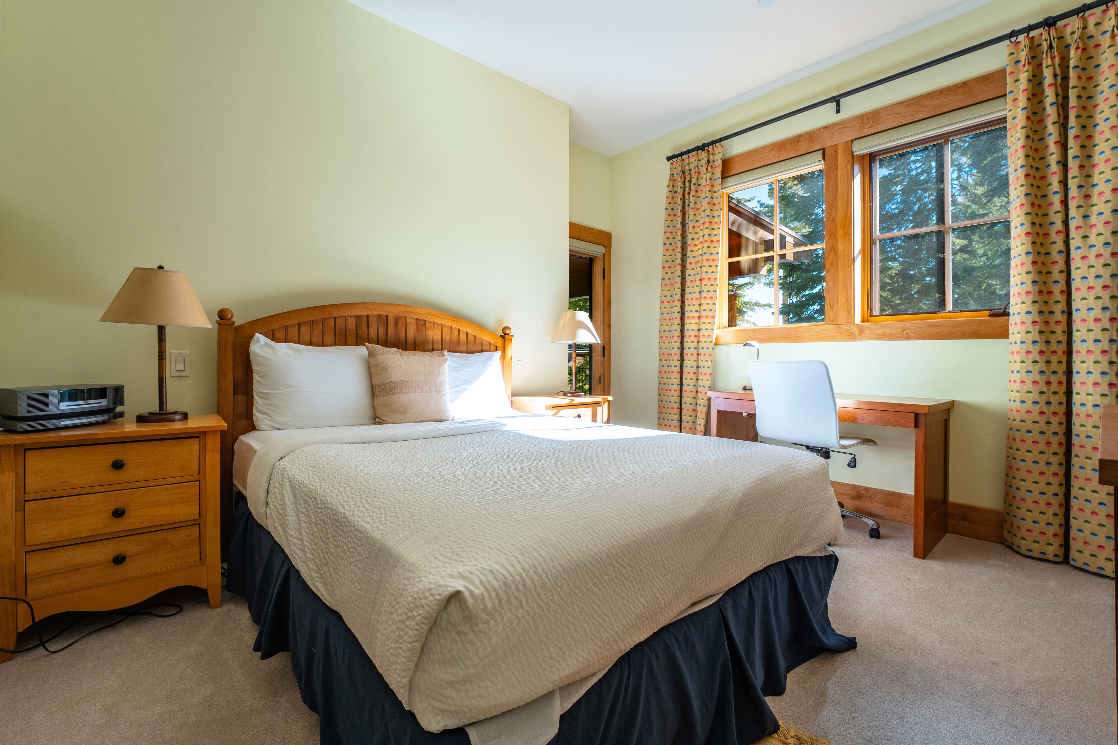 This is a cozy guest bedroom featuring a wooden-framed bed with neutral-toned bedding and a dark blue bed skirt. A wooden nightstand with a lamp sits beside the bed, and a desk with a white chair is positioned near the window, which is adorned with patterned curtains. The room has a warm and inviting atmosphere, perfect for a comfortable stay.