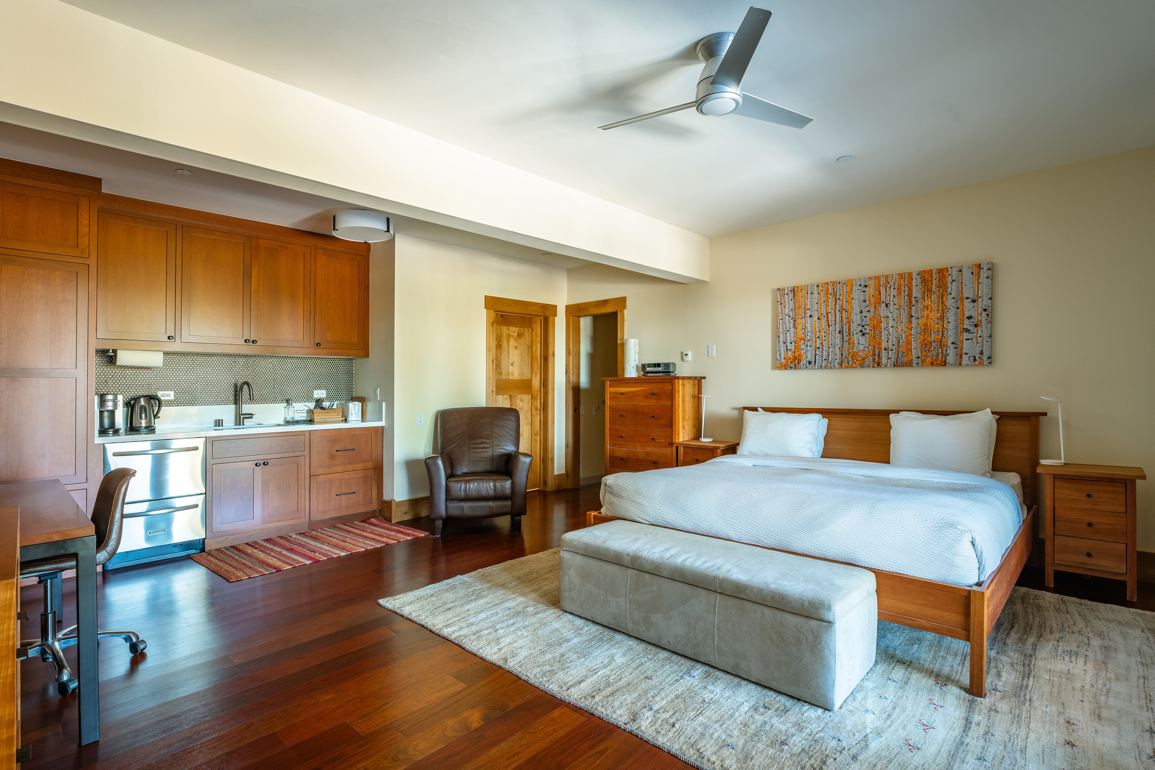 This is a well-appointed primary bedroom featuring a king-size bed with a light-colored duvet, flanked by wooden nightstands. A seating bench is placed at the foot of the bed, and a piece of artwork hangs above the headboard. The room also includes a kitchenette with wooden cabinets and stainless steel appliances, along with a leather armchair, creating a comfortable and functional space.