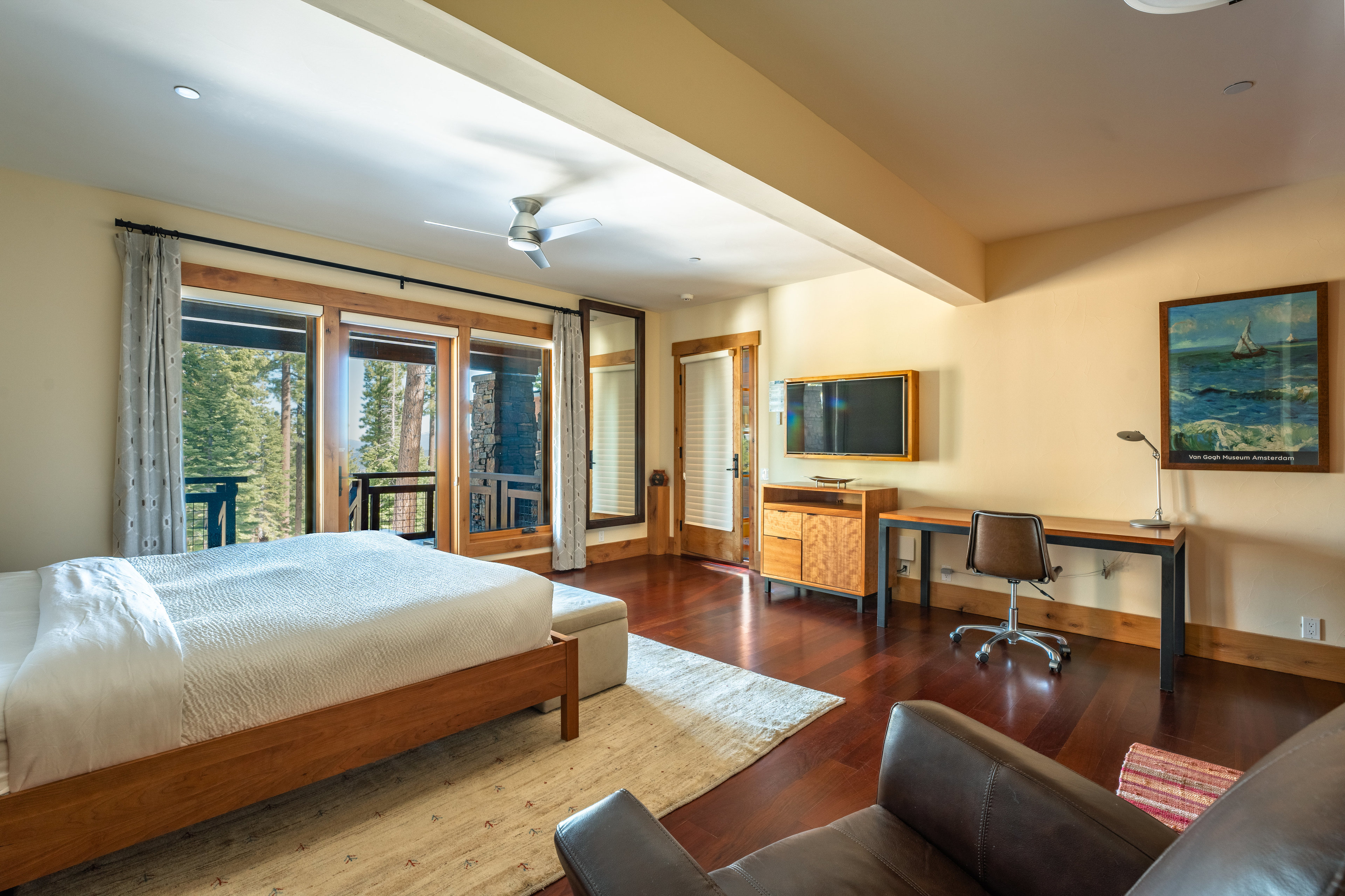 This is a primary bedroom featuring a wooden bed frame with a white comforter, a light-colored rug, and a desk area with a chair. Large windows offer natural light and views of the surrounding trees, while a TV is mounted on the wall above a wooden cabinet. The room has a warm and inviting atmosphere with hardwood floors and neutral wall colors.