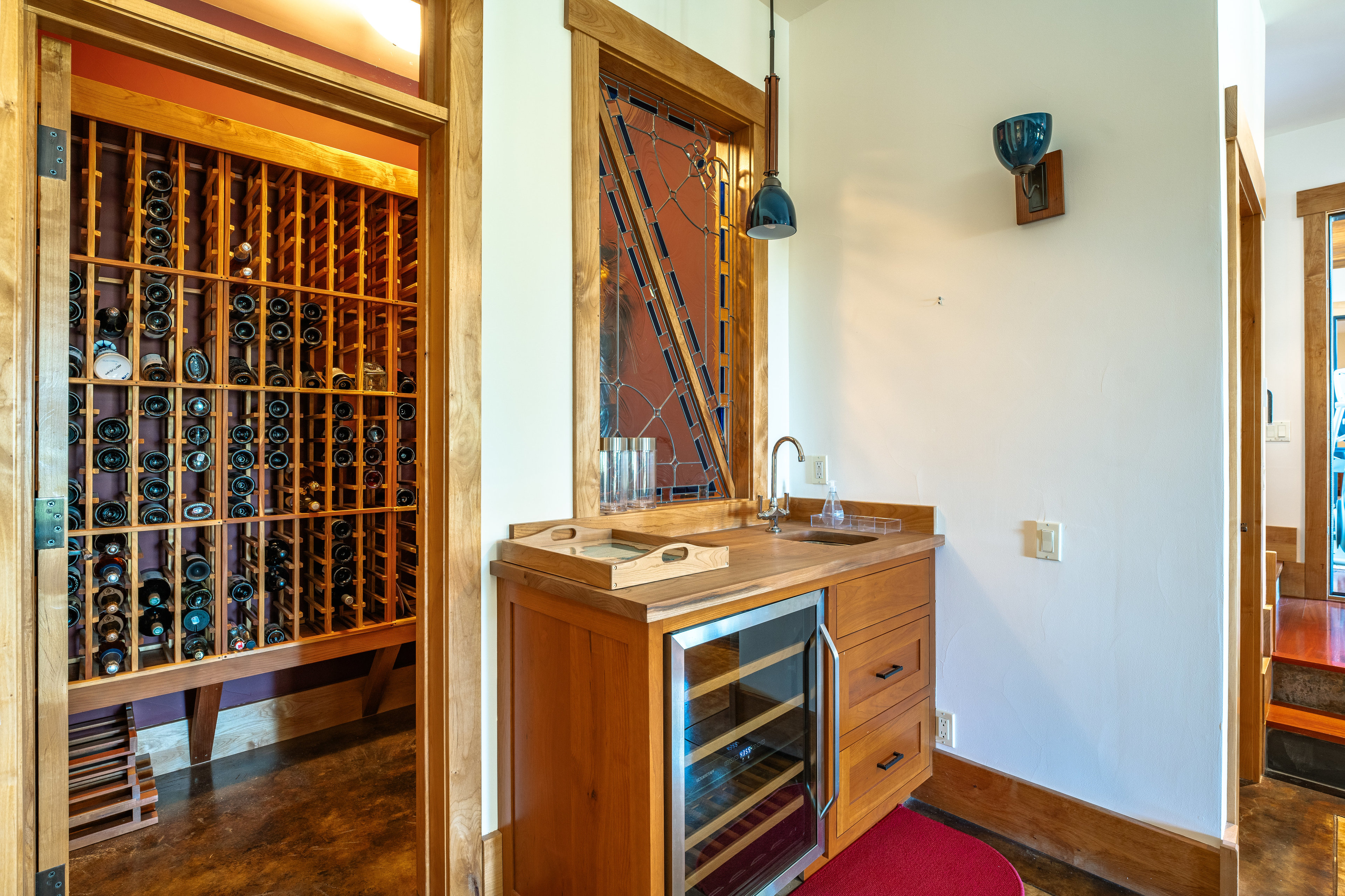 This interior shot showcases a well-organized wine cellar adjacent to a stylish wet bar. The wine cellar features wooden racks filled with bottles, while the wet bar includes a sink, countertop, and a built-in wine refrigerator. The space is illuminated by a pendant light and a decorative wall sconce, creating an inviting atmosphere for wine enthusiasts.