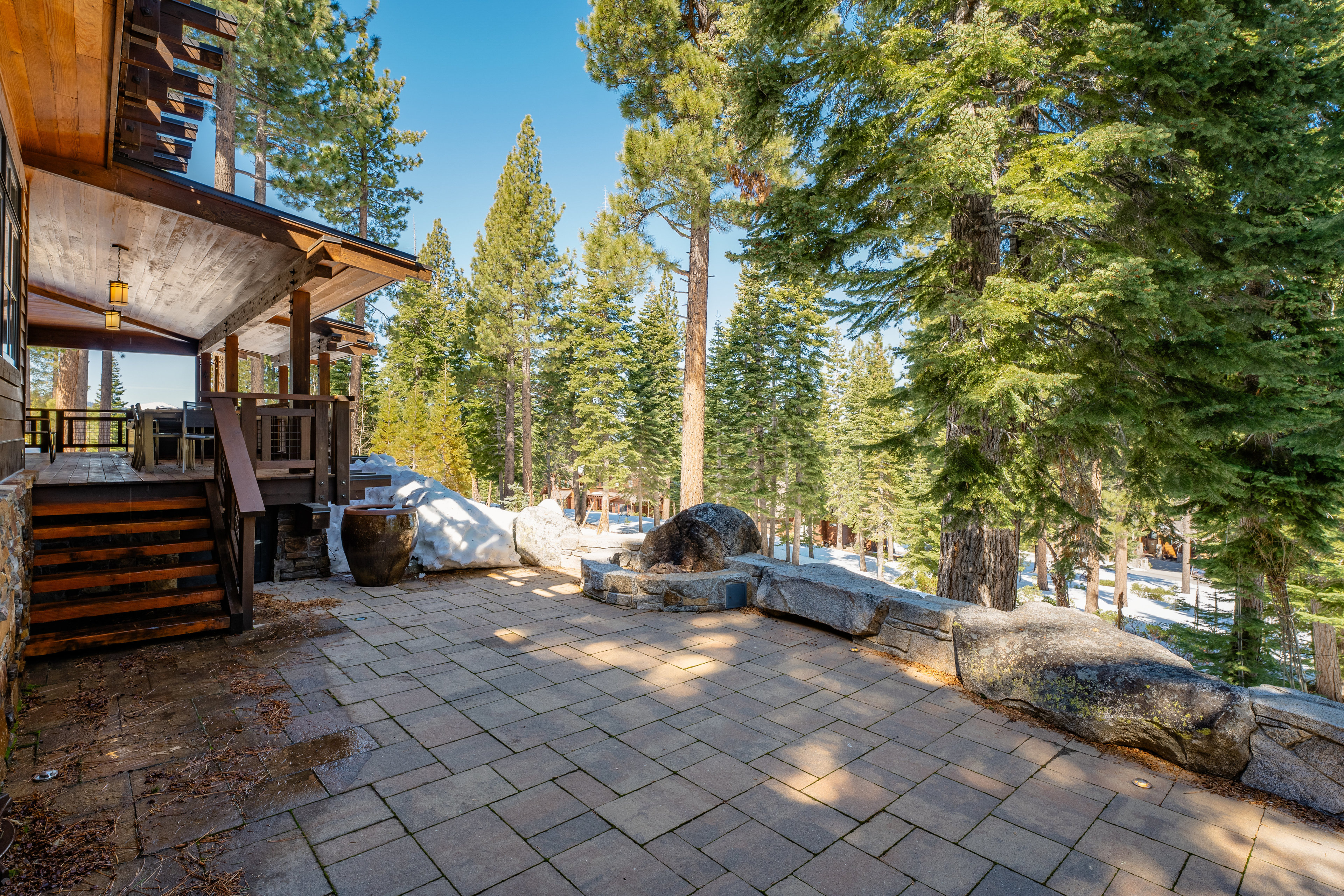 This image showcases a spacious outdoor patio area, constructed with stone pavers and bordered by a natural stone retaining wall. The patio is adjacent to a wooden deck with stairs leading down, and the surrounding landscape features mature pine trees and patches of snow, creating a serene and private setting. The area appears well-maintained and designed for outdoor living and entertaining.