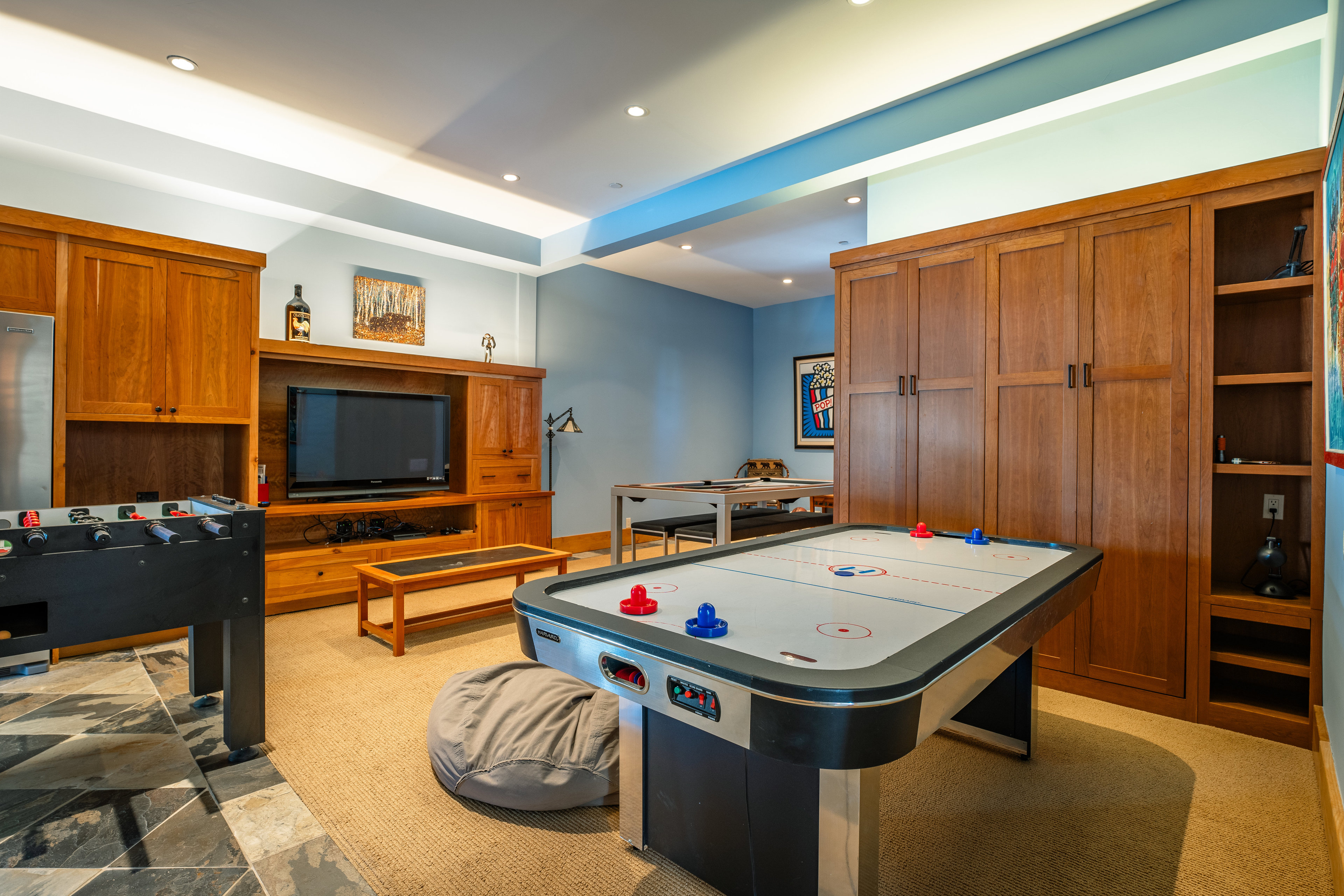 This is a well-appointed basement featuring an air hockey table, foosball table, and a pool table in the background. The room is decorated with wooden cabinetry and a light blue wall color, creating a recreational space. The flooring transitions from carpet to tile, adding visual interest.