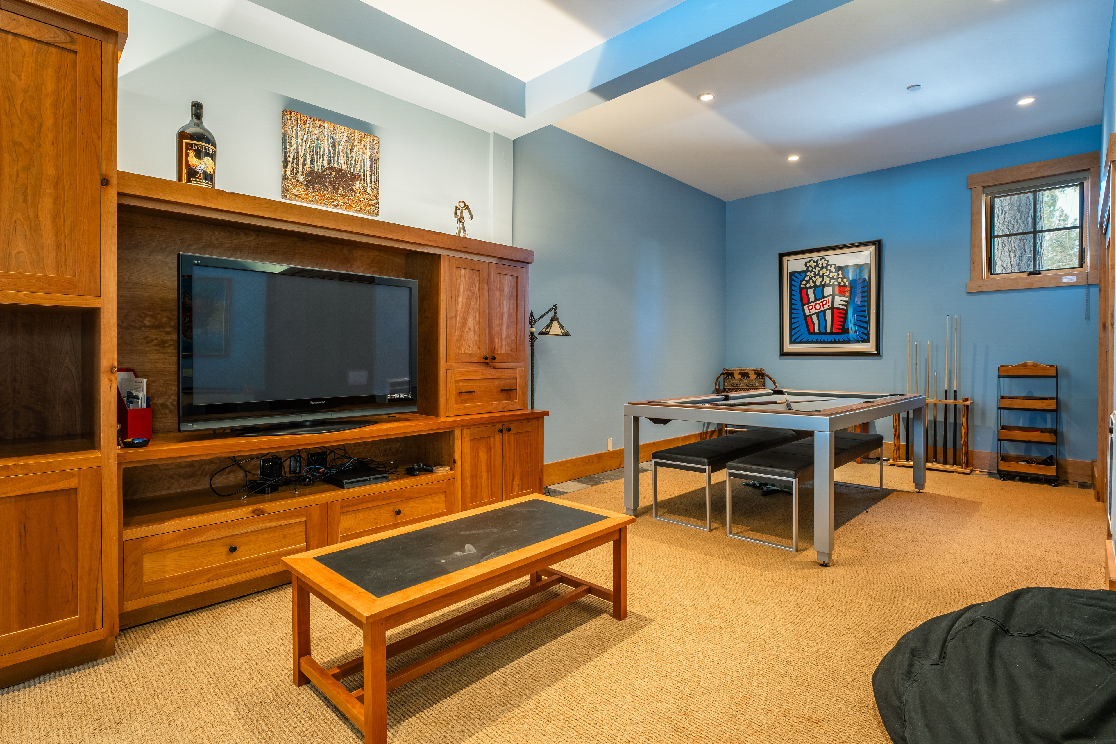 This interior shot showcases a living room with a pool table, a large wooden entertainment center with a flat-screen TV, and a coffee table. The walls are painted a calming blue, and the room is carpeted in a neutral tone. The space appears to be designed for relaxation and entertainment.