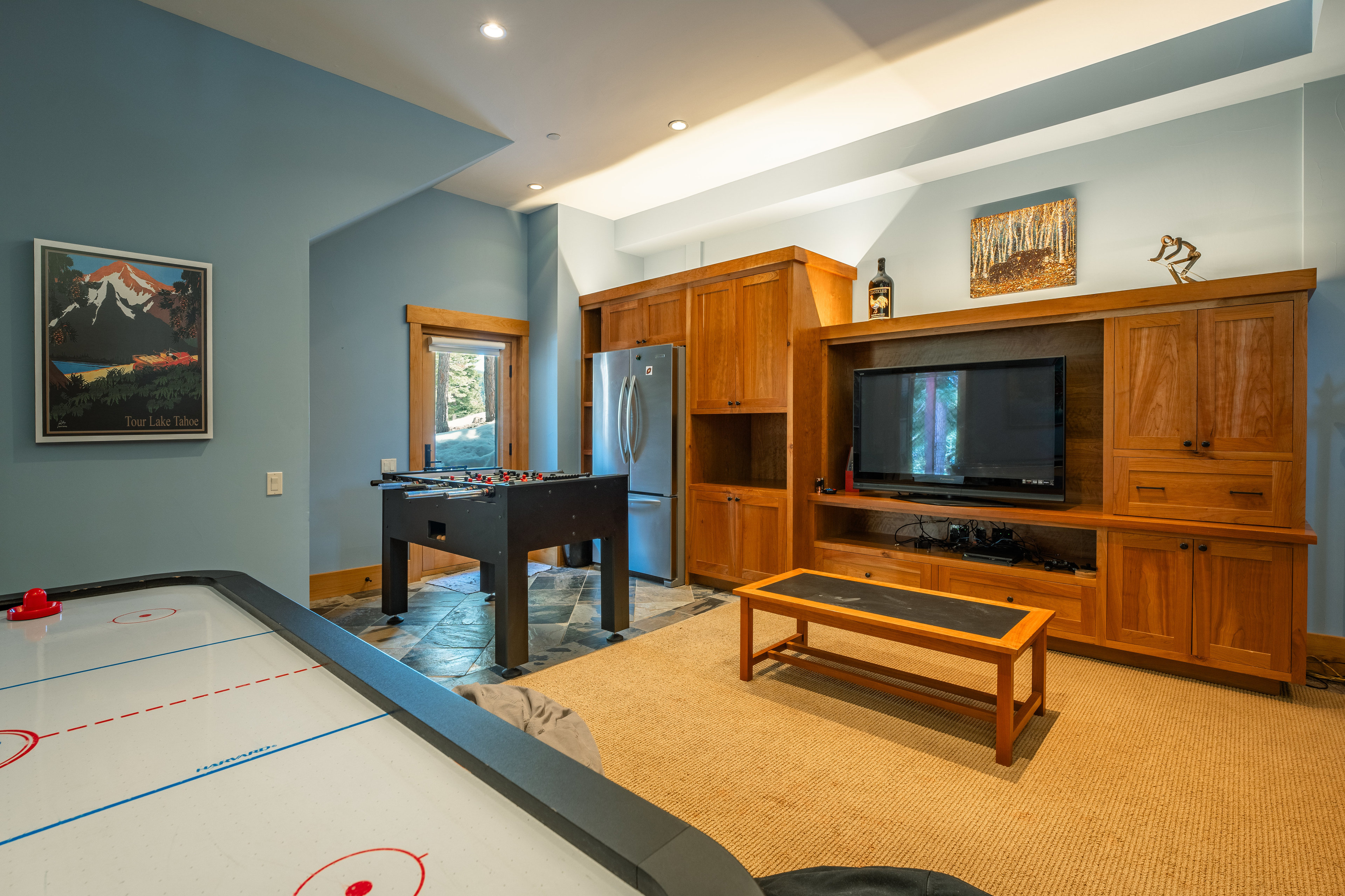This interior shot showcases a finished basement area designed for entertainment. The room features a foosball table, an air hockey table, and a large wooden entertainment center with a television. A small wooden coffee table sits in front of the entertainment center, and the room is carpeted, creating a cozy and inviting atmosphere.