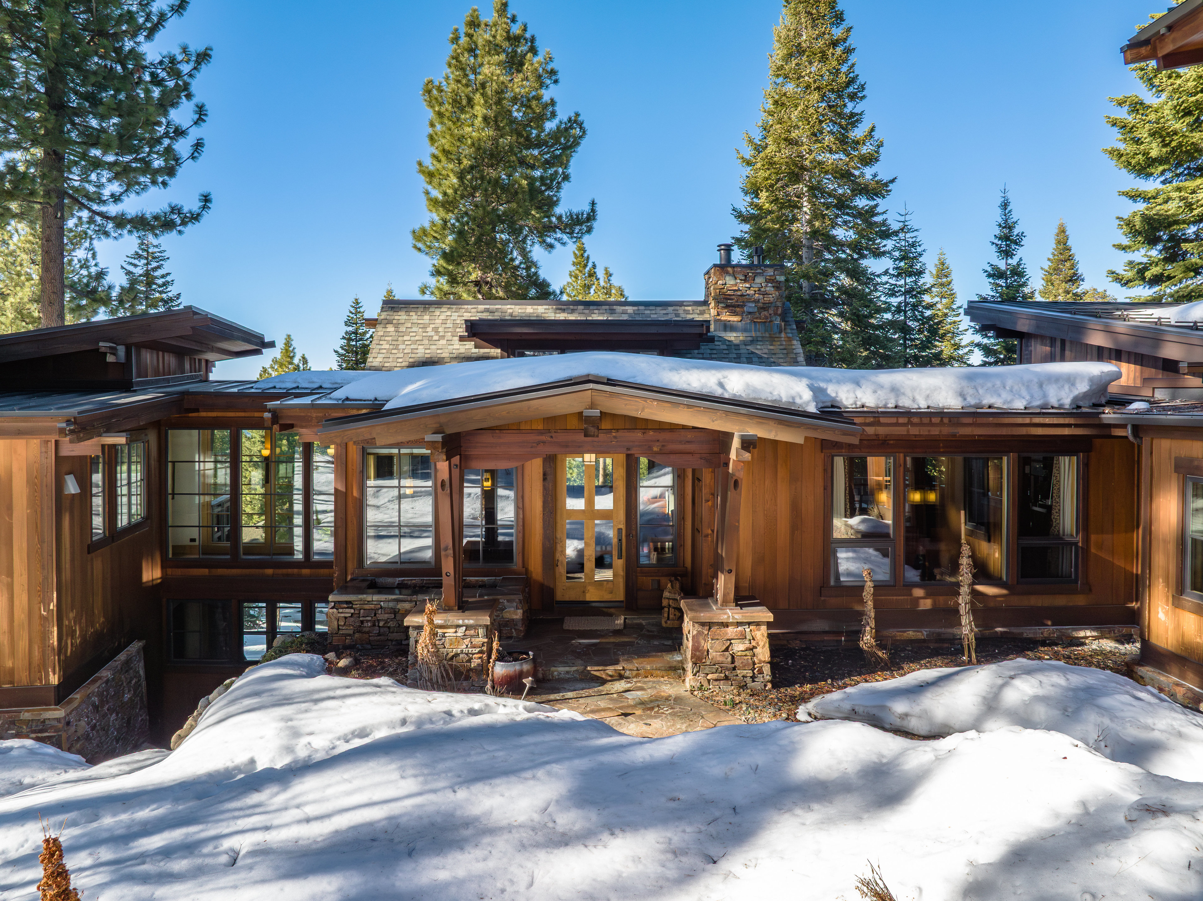 This is a front exterior shot of a wood-clad home with a stone and wood entryway. Snow covers the ground, and tall evergreen trees surround the house. The home features large windows and a stone chimney, giving it a rustic yet modern feel.