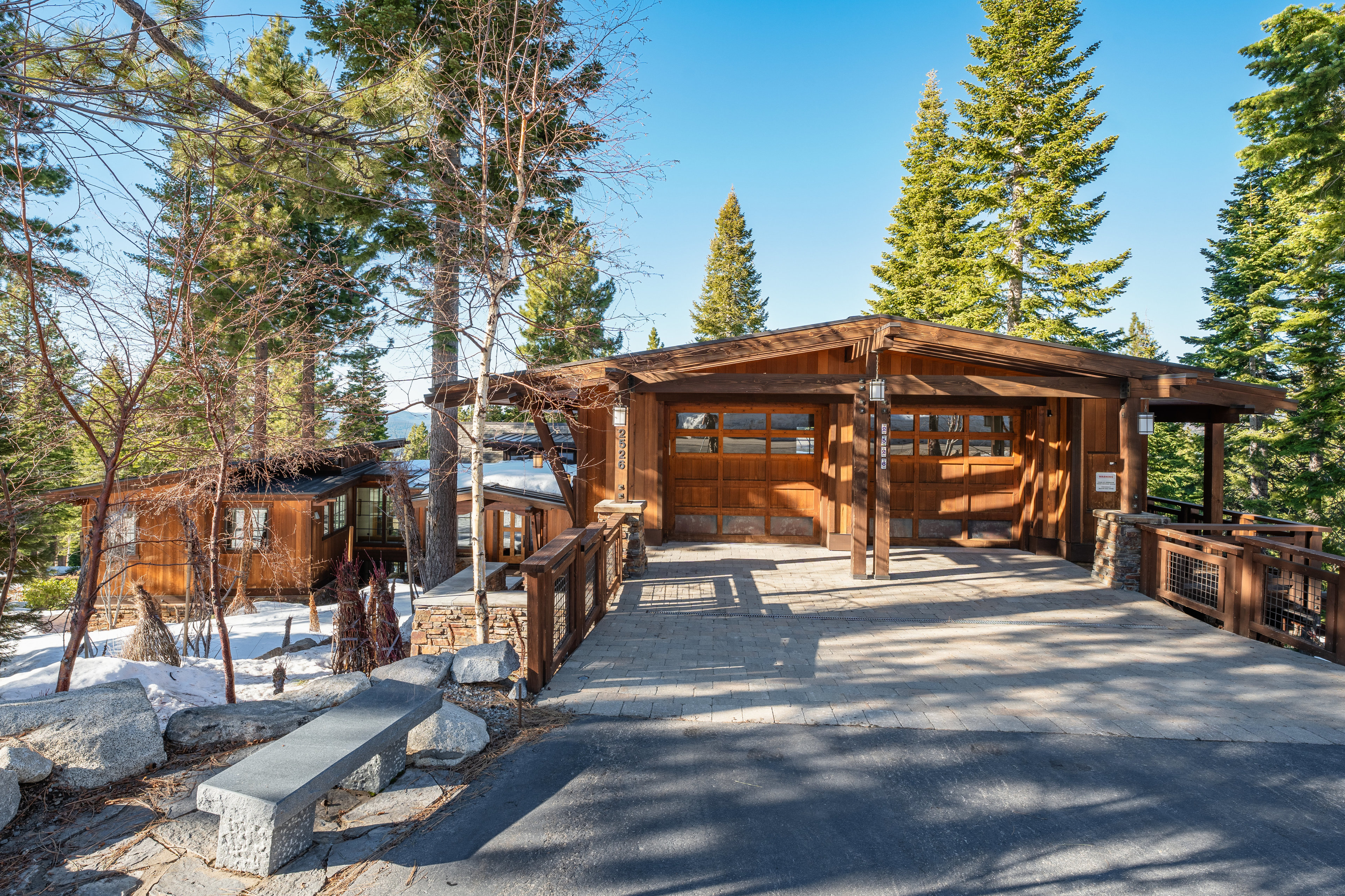 This image showcases a charming wooden garage with two bays, connected to the main house by a stone-paved walkway. The structure is surrounded by tall pine trees and features a stone foundation, blending seamlessly with the natural landscape. The driveway leads up to the garage, offering a welcoming entrance to the property.
