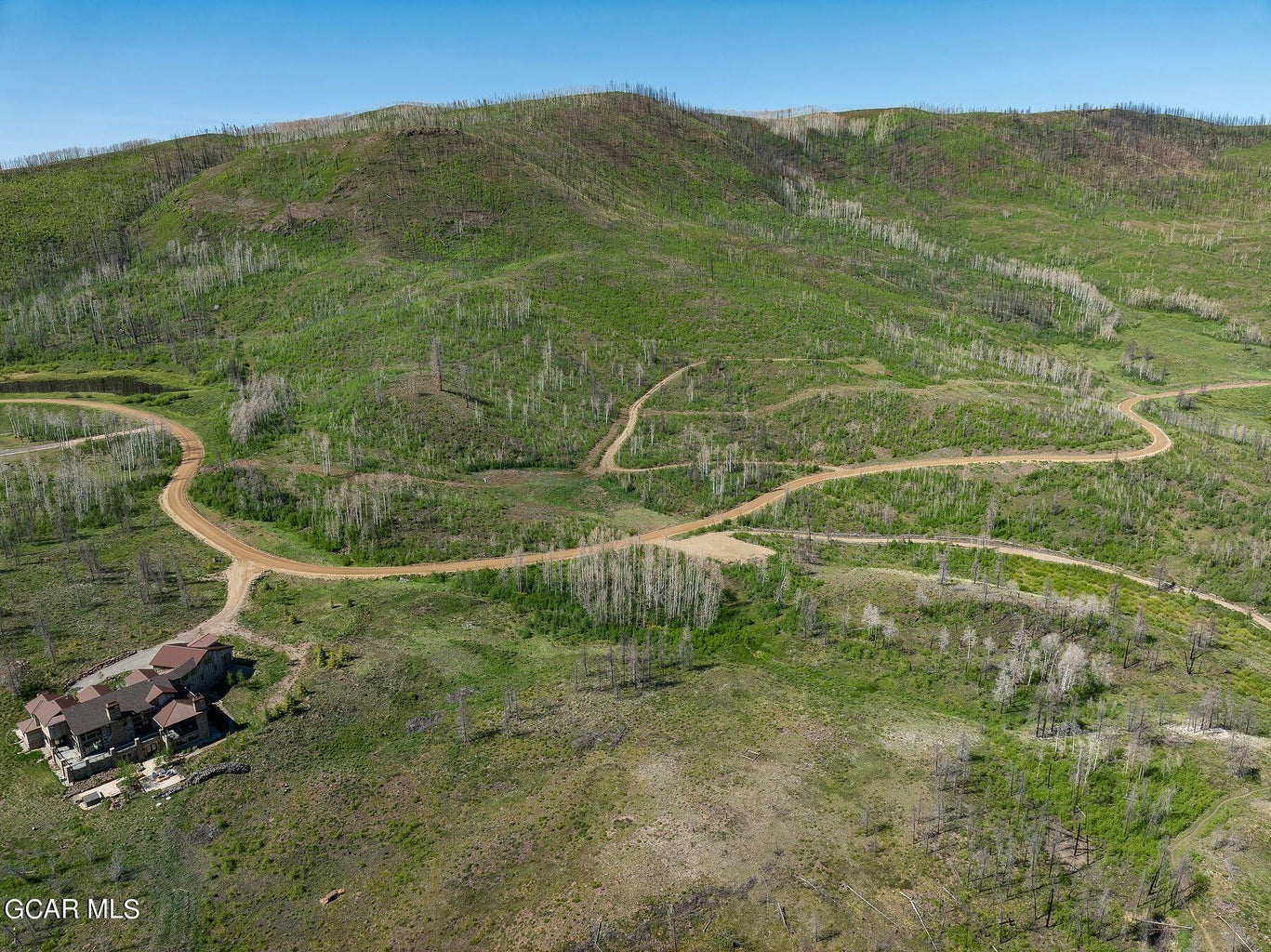 This high-altitude aerial view showcases a large, custom residence nestled within a vast, hilly landscape. The property features extensive natural terrain, burnt timber, and a winding dirt access road that traverses the expansive hillside. This location offers significant privacy and scenic potential, typical of rural mountain estate properties.
