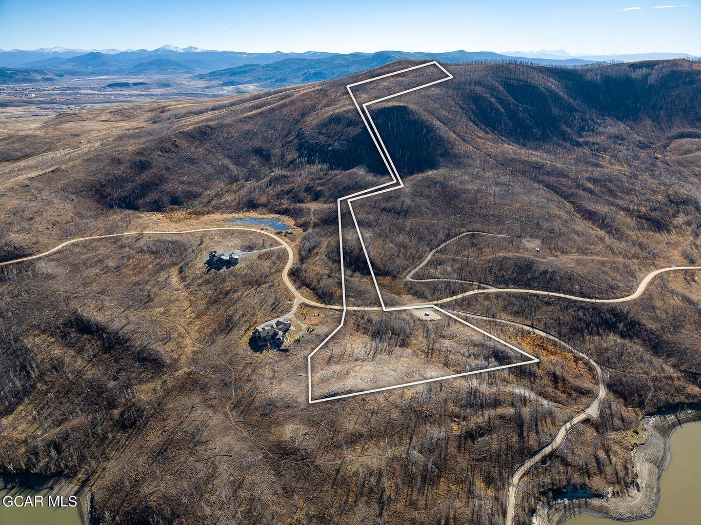 This is an expansive aerial view showing a large parcel of rugged, mountainous land highlighted by a white outline. The property features hilly terrain with sparse vegetation and a gravel access road that winds towards existing residences nearby. It offers a remote, rural setting with scenic landscape vistas suitable for custom development.