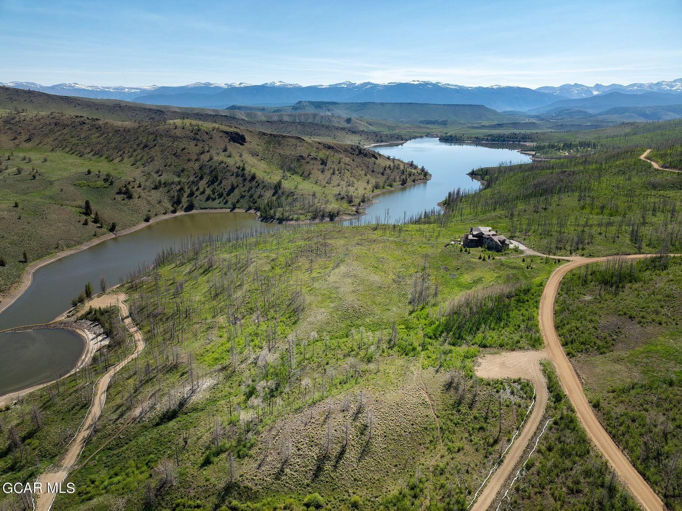 This stunning wide-angle aerial shot captures a luxury home situated on a sprawling, rugged hillside overlooking a large reservoir. The property features a naturalistic landscape with mature and re-growing vegetation, extensive private dirt roads, and panoramic mountain views in the background. It offers a remote, serene setting ideal for those seeking privacy and breathtaking natural vistas.
