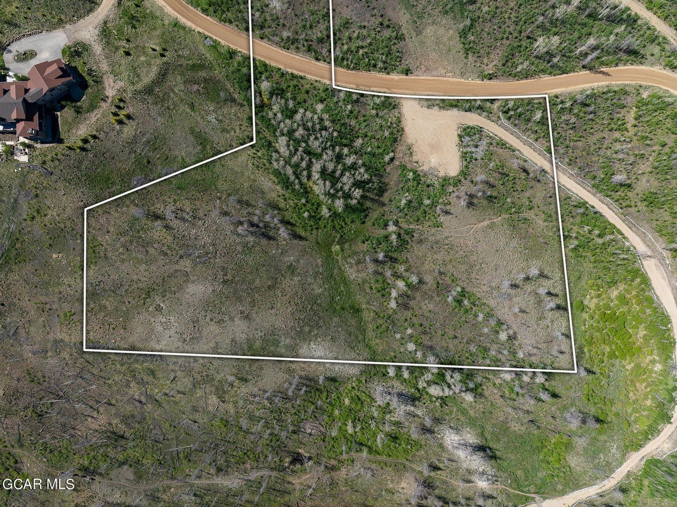 This is an aerial, top-down perspective of a large, vacant plot of land highlighted by a white boundary outline. The terrain is characterized by a mix of open, grassy areas, pockets of trees, and a dirt driveway providing access. It offers a clear view of the property's size and its proximity to established neighboring structures and surrounding wilderness.