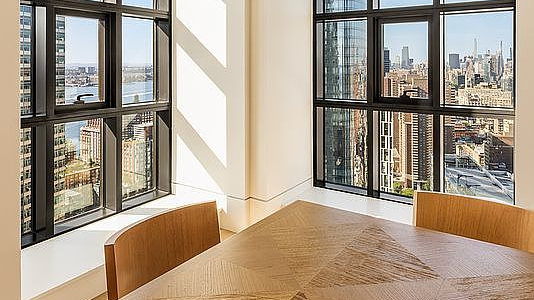 This bright and sophisticated dining area features a warm wood-toned table with a distinct geometric pattern. The room is framed by large, multi-pane windows that provide an expansive, panoramic view of the cityscape. The minimalist design and natural light create a clean, modern, and high-end atmosphere ideal for urban living.