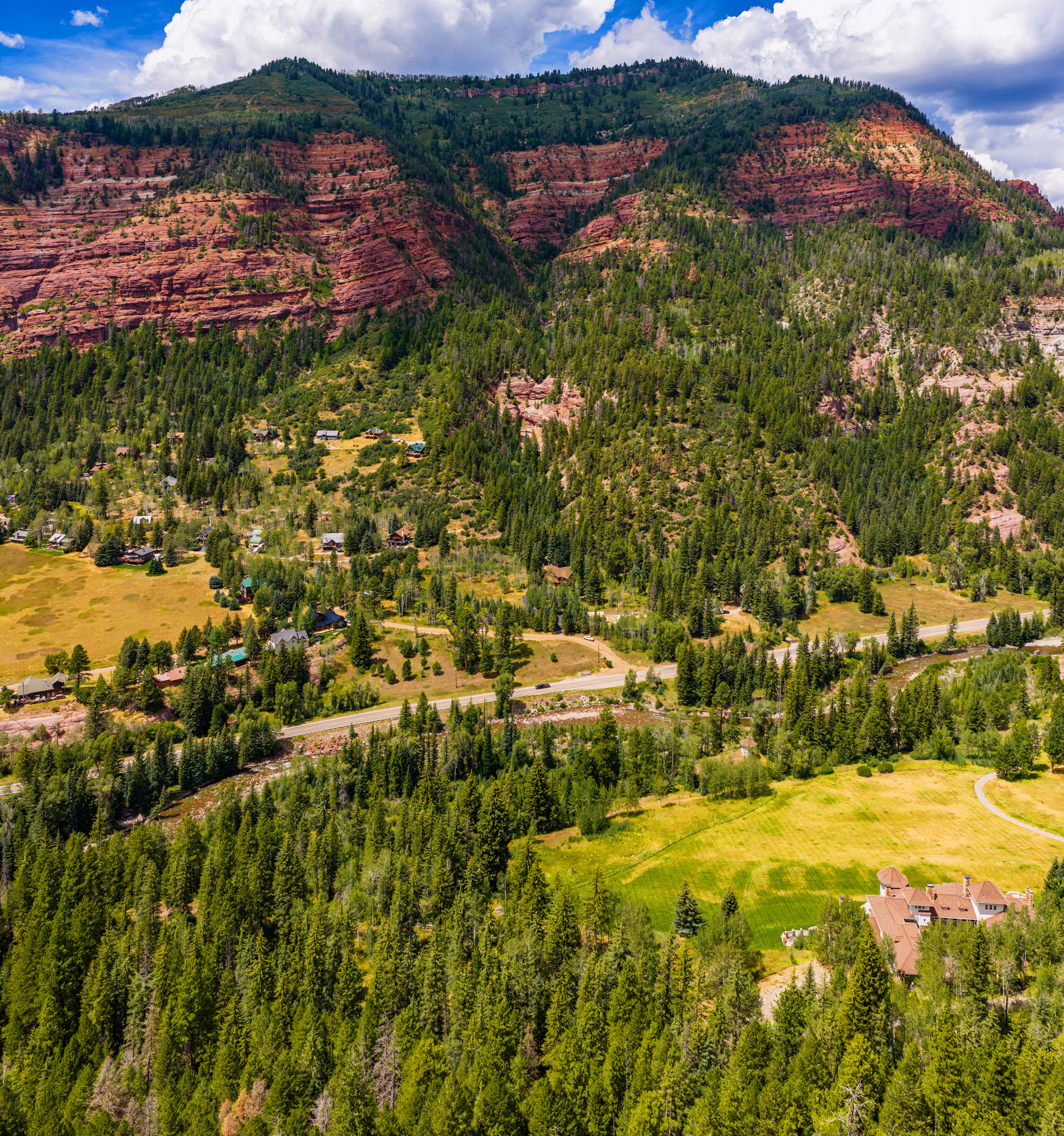 This expansive aerial view captures a sprawling mountain estate nestled within a lush, forested valley beneath dramatic red rock cliffs. The property features a large, multi-gabled residence situated on a manicured lawn, with a winding path leading through the grounds. A river meanders through the landscape, paralleled by a road that connects the home to a small, secluded residential community tucked into the hillside.