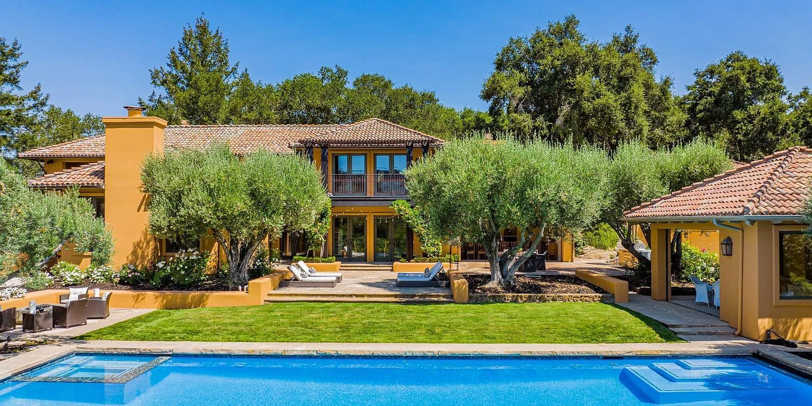 An ochre-colored estate and swimming pool framed by olive trees
